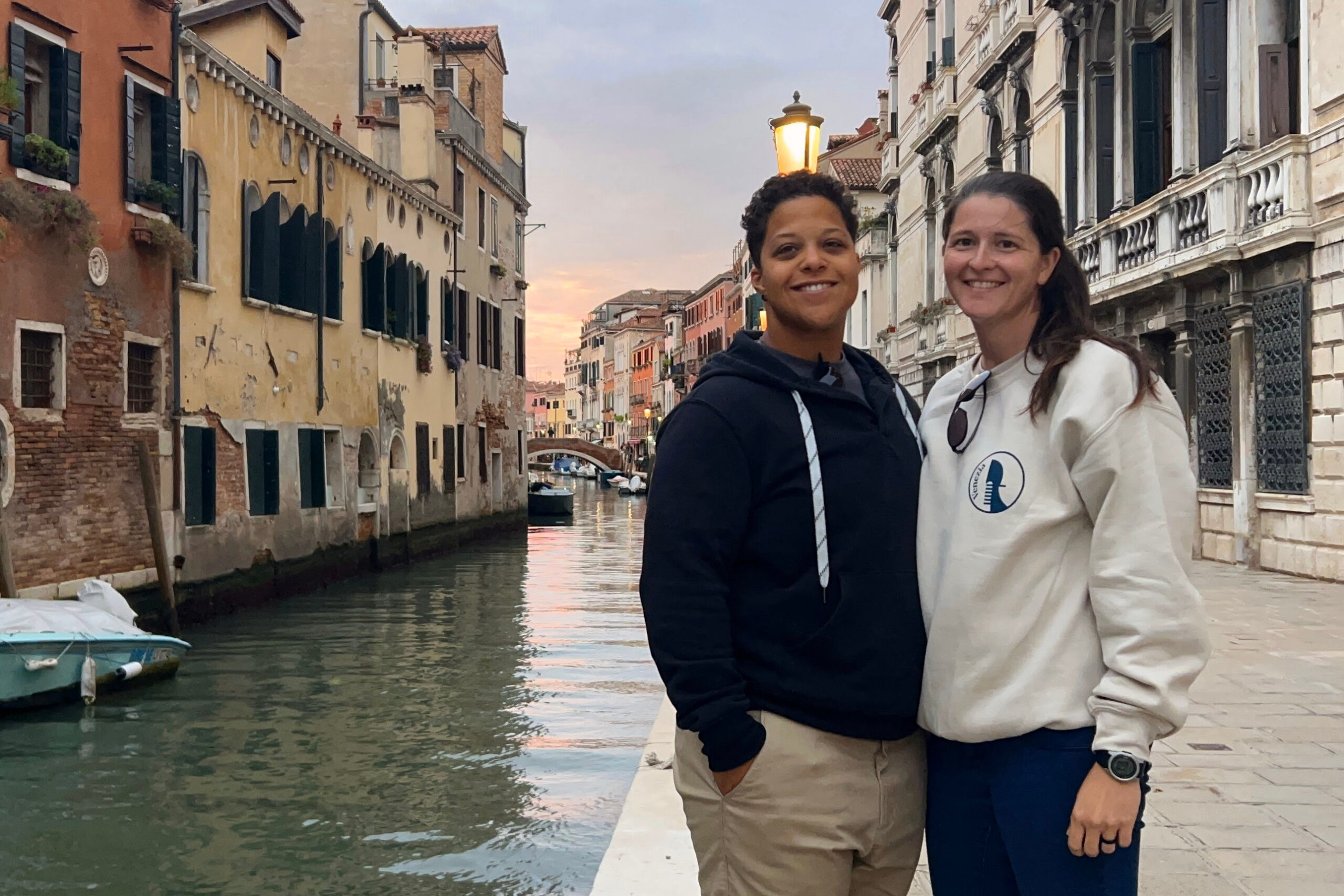 Two people stand close together by a canal in a sunny Venetian street, smiling at the camera, with pastel buildings lining the waterway behind them.