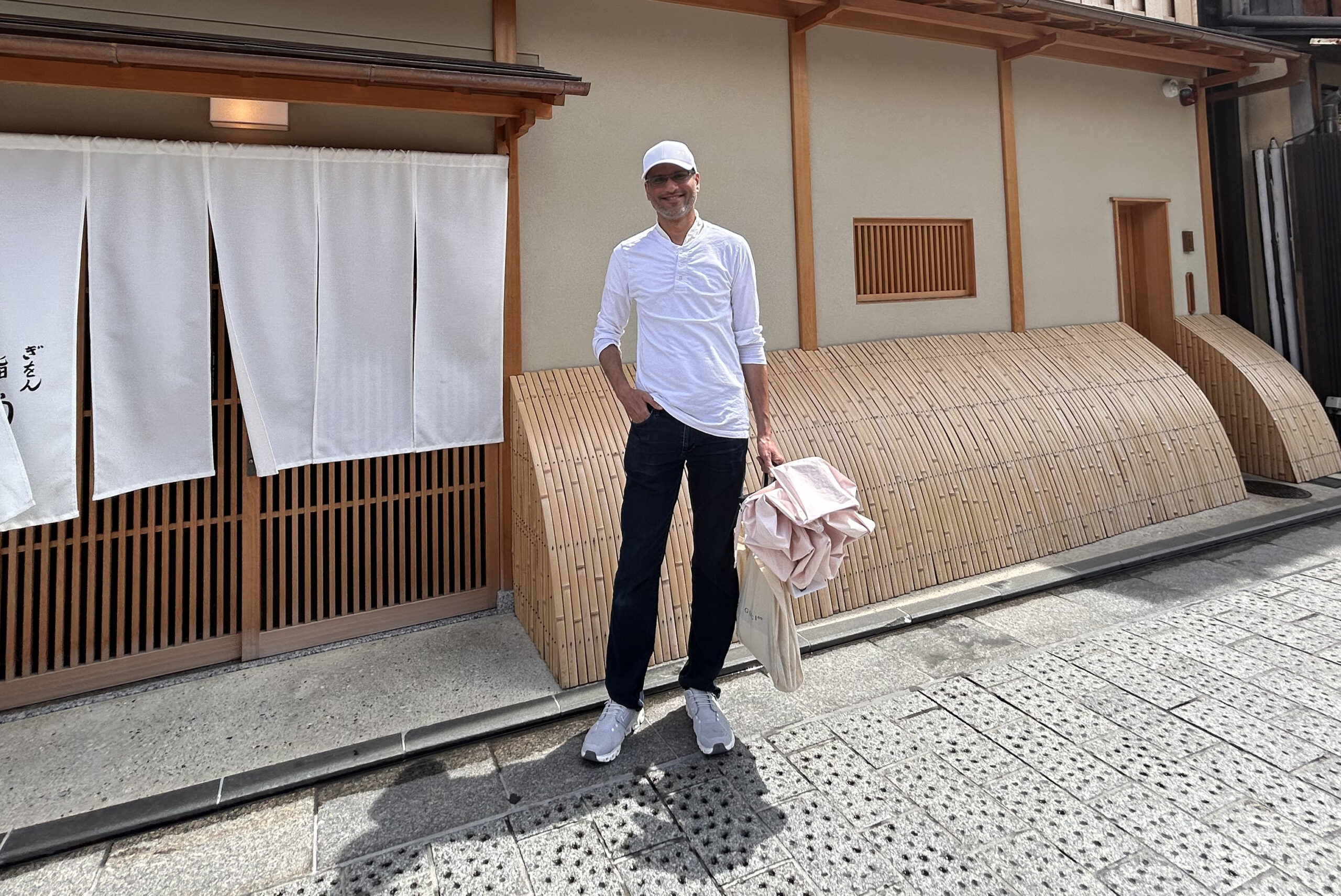 Smiling man in a white shirt and cap stands on a sidewalk in front of a Japanese-style storefront with white curtains and wooden latticework, carrying a beige tote bag with pink fabric piece inside.
