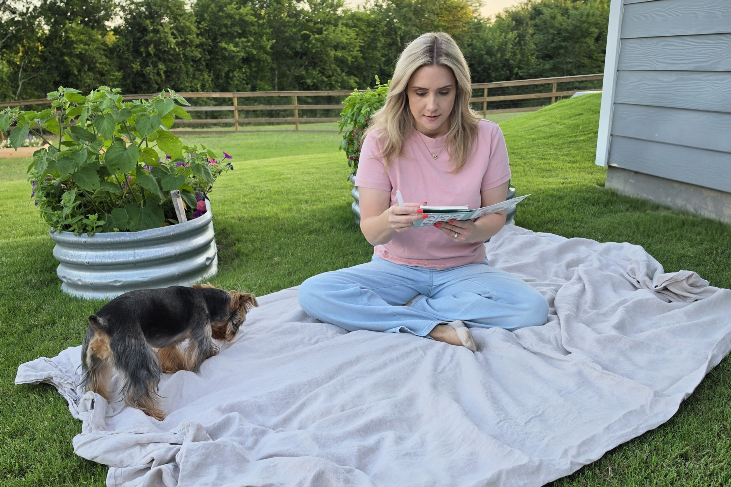 Woman in a pink shirt sits on a blanket reading a magazine outdoors, with a small dog nearby in a grassy backyard.