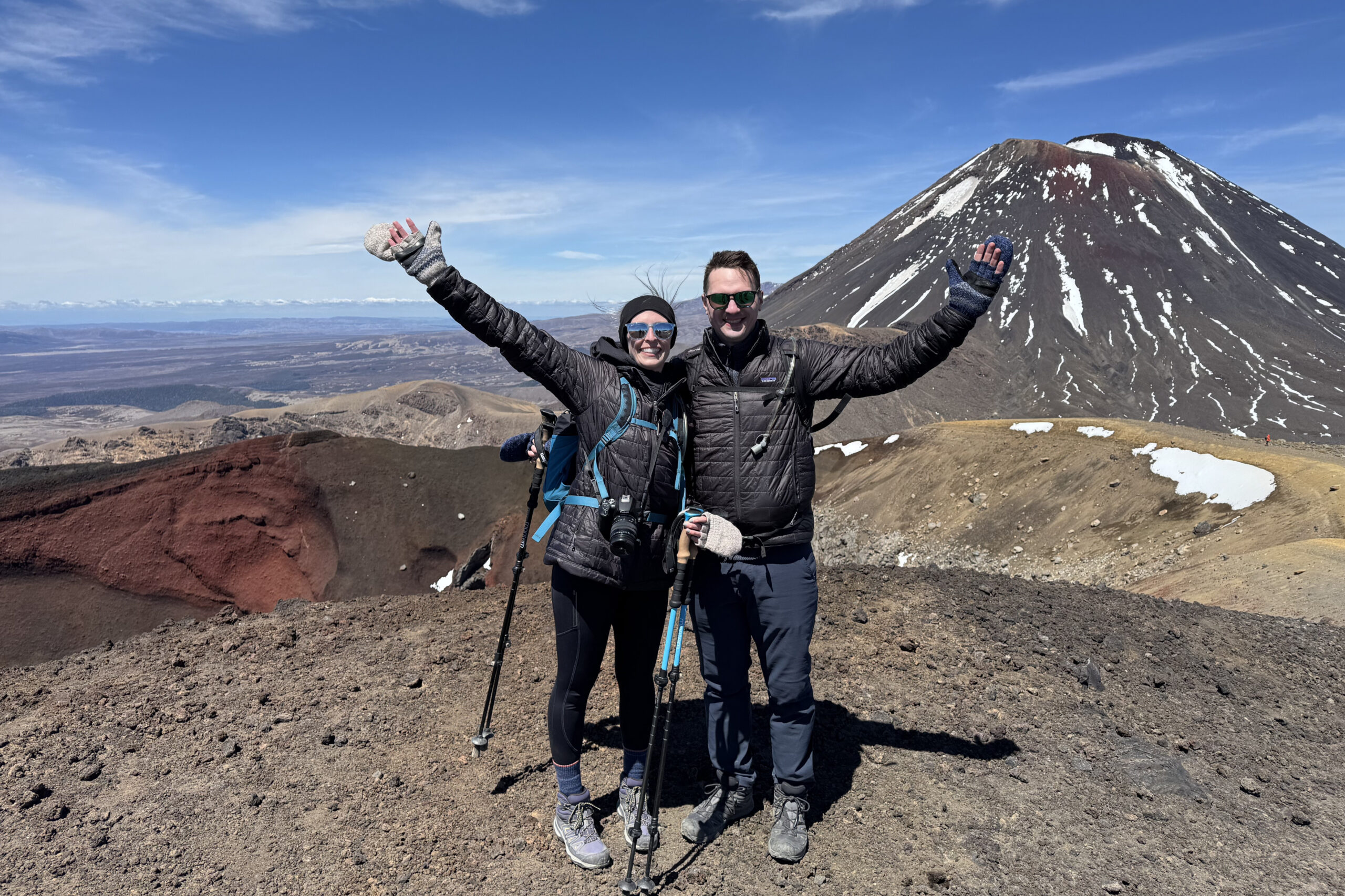 Two hikers in winter gear pose with arms raised on a volcanic landscape, a snow-dusted cone behind them under a clear blue sky.