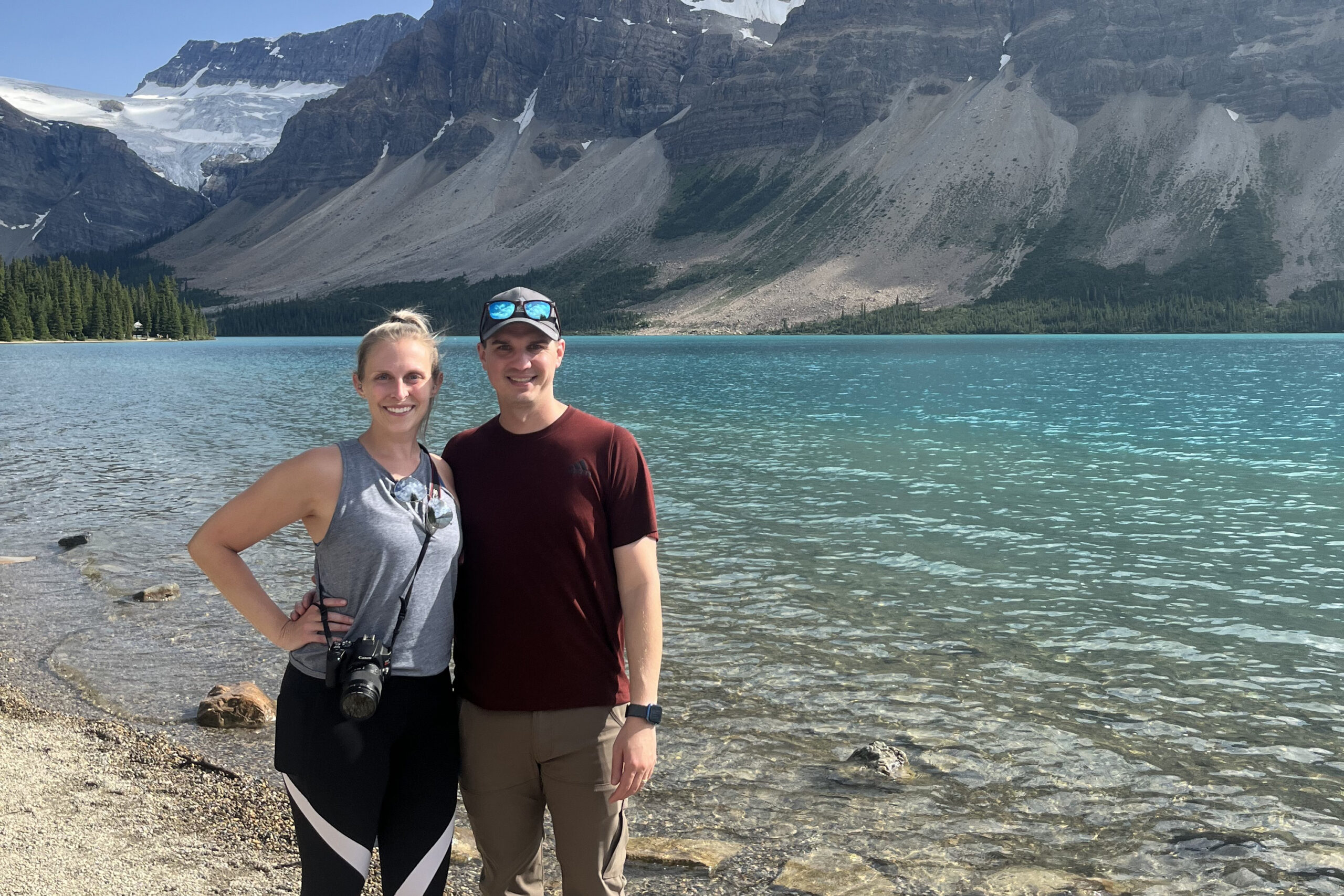 Smiling couple posing by a turquoise alpine lake with rocky mountains and evergreen forest in the background.