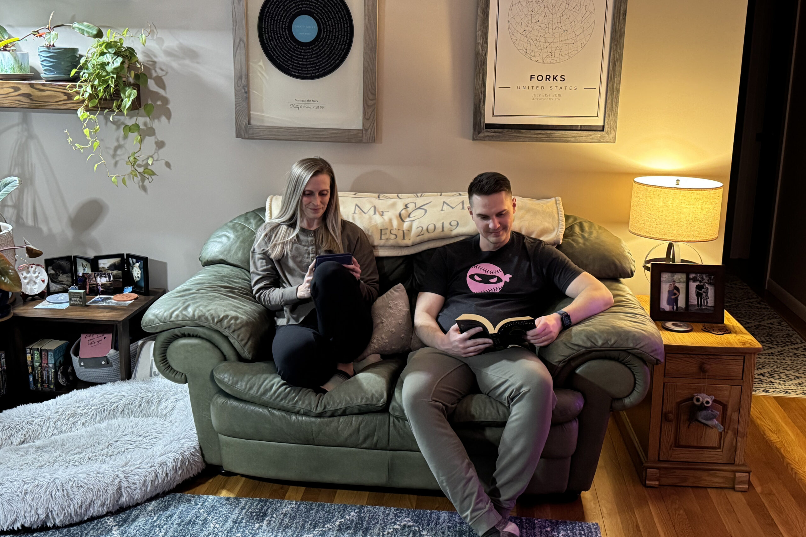 Two people sit on a green sofa in a cozy living room; woman uses a phone, man reads a book beside her.