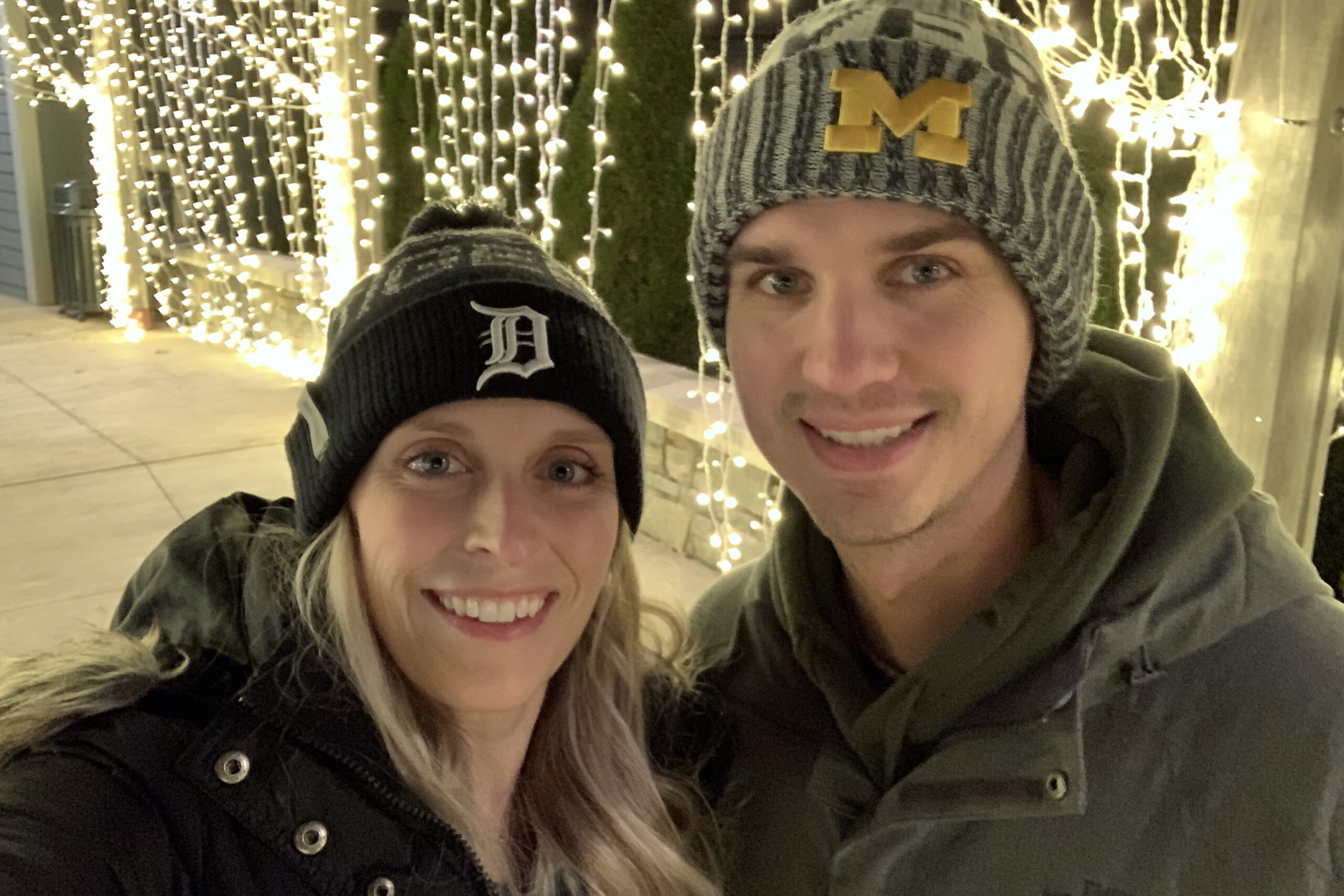 Close-up selfie of a smiling couple wearing winter hats, with string lights in the background at night.