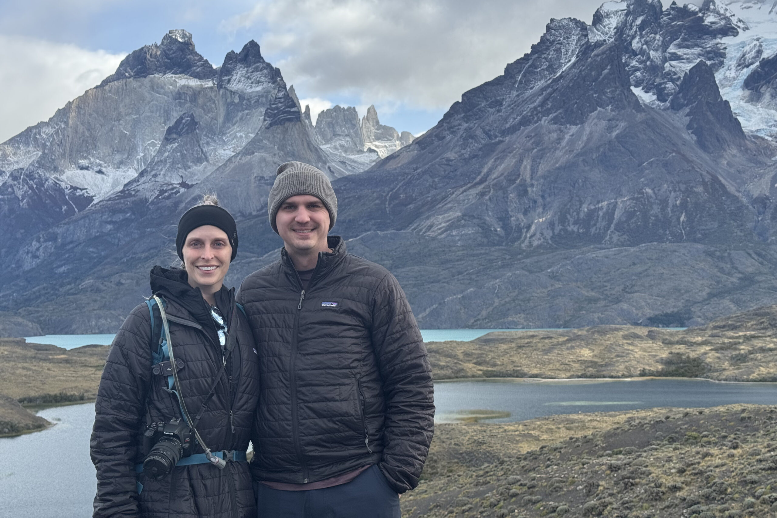 Two hikers in warm jackets pose for a photo with rugged mountains and a lake in the background, cloudy sky above.