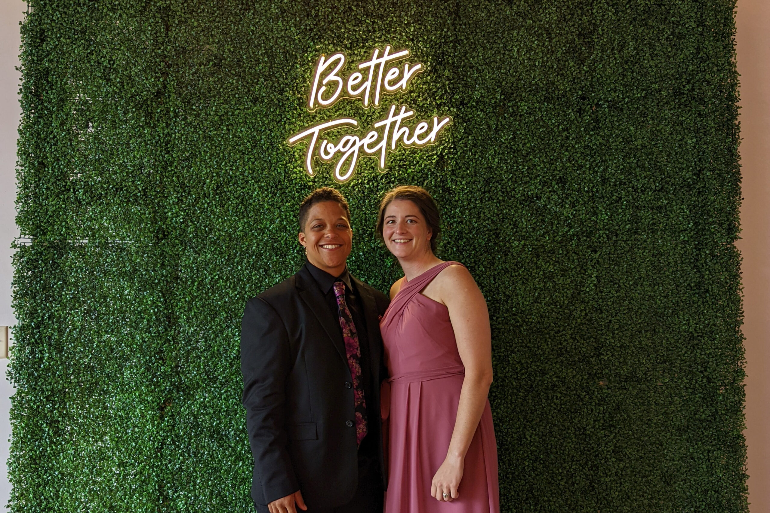Two smiling adults in formal attire posing in front of a green hedge wall with a neon 'Better Together' sign above them.