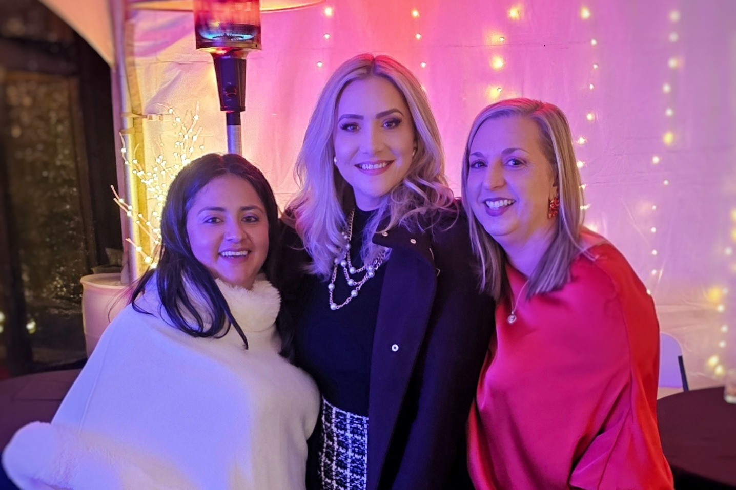 Three women smiling and posing together at a festive event under pink-purple lighting with string lights behind them.