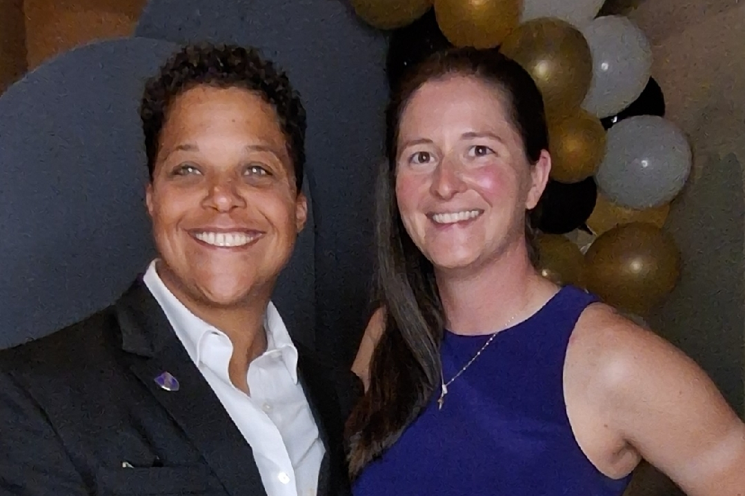 Two women posing and smiling at a celebration; one in a black blazer with a white shirt and the other in a royal blue sleeveless dress, with gold and white balloons in the background.