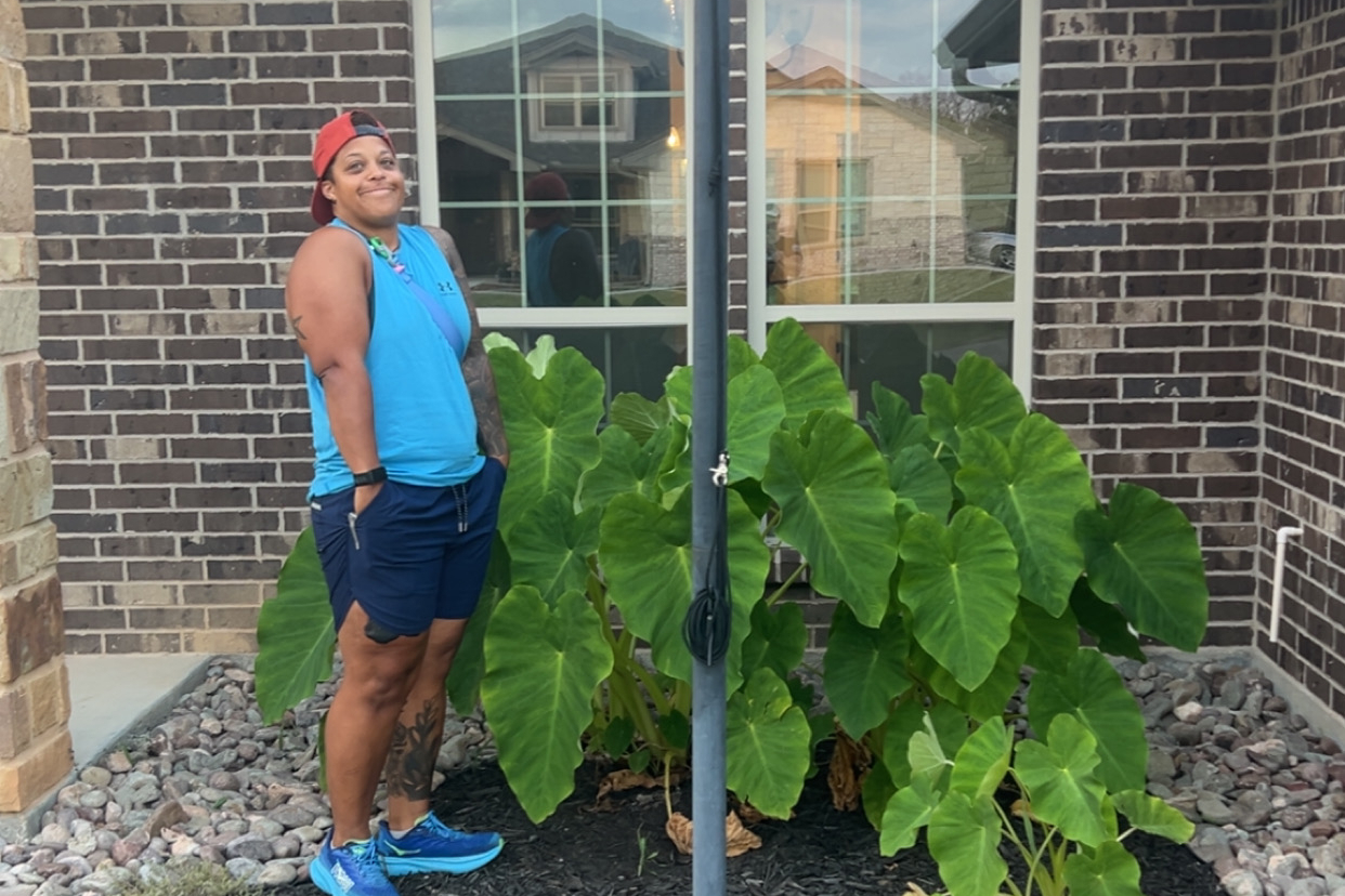 A person in a blue tank top and shorts stands with hands in pockets beside large green elephant ear plants by a brick house front.