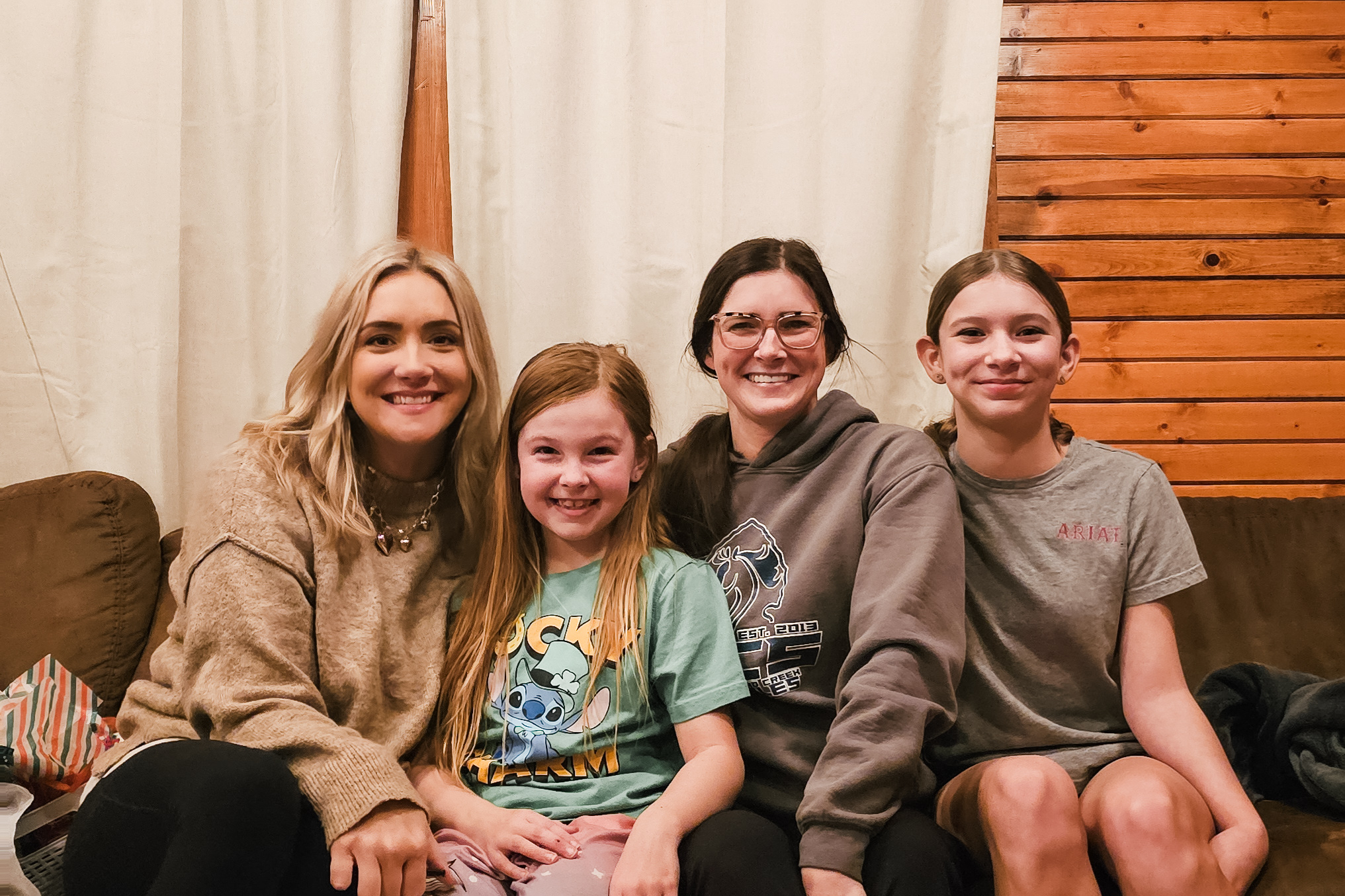 Four smiling people sit close together on a couch indoors, with beige curtains behind and a wooden wall to the right.