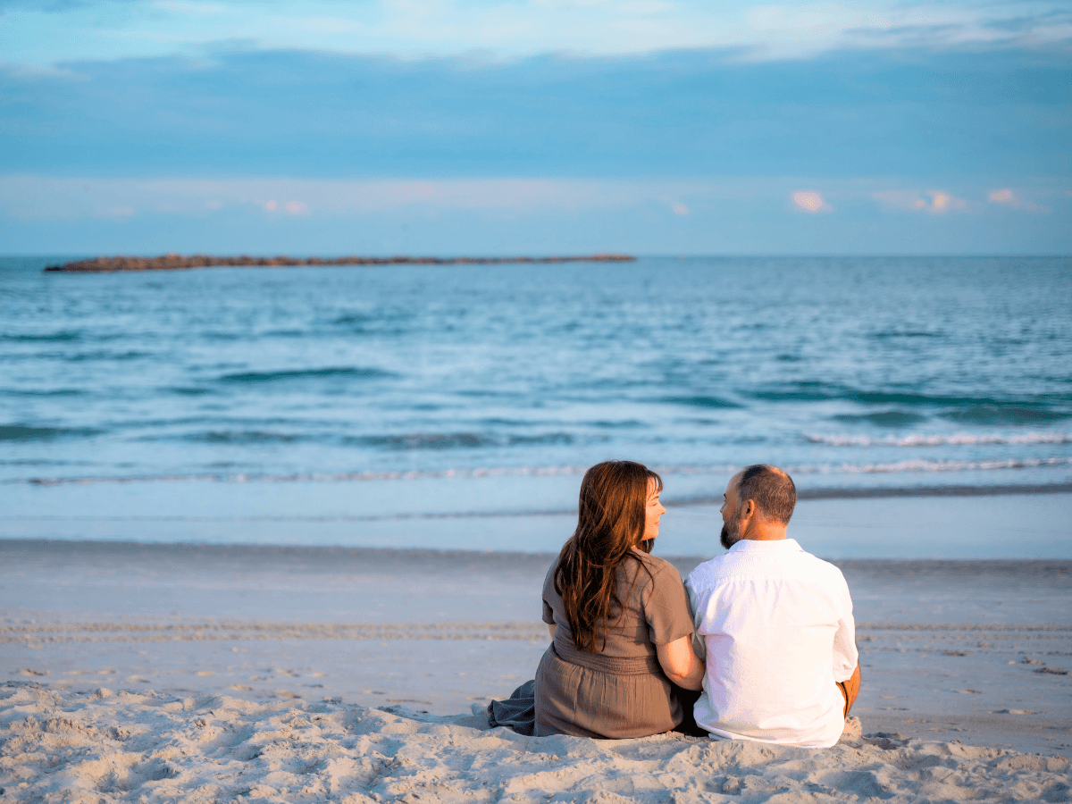 Couple sits on a sandy beach, facing each other and talking as waves roll in behind them.