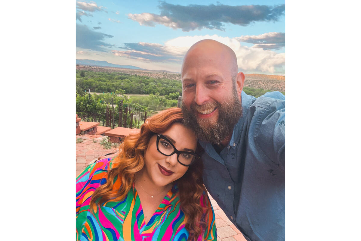 Smiling couple taking a selfie on a sunny terrace with a green valley and blue sky in the background.