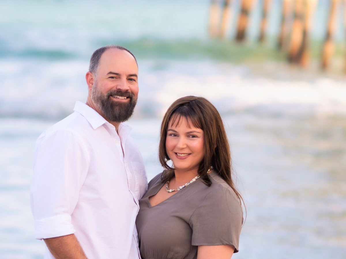 Smiling couple standing on a sunny beach with waves and a pier in the background, near the shoreline.