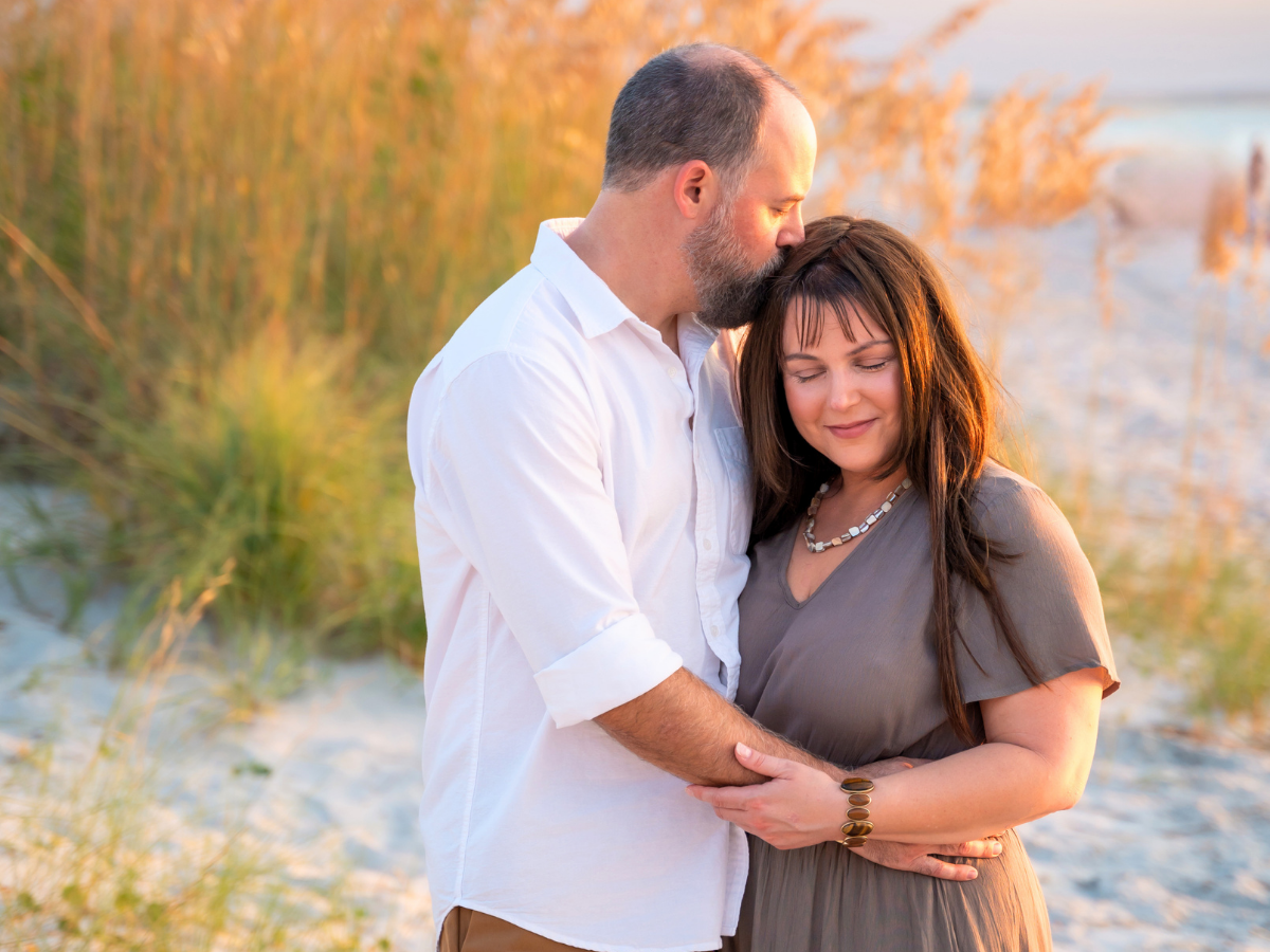 Couple embracing on a sandy beach at sunset, man kissing the woman's head as they hold hands.