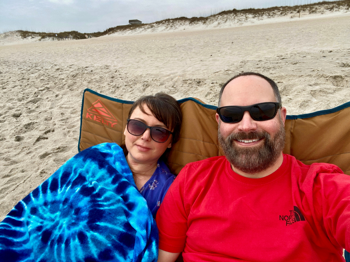 Couple wearing sunglasses sits in a beach chair on light sand with a blue tie-dye blanket, smiling at the camera under a cloudy sky.