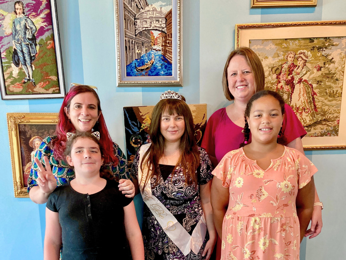 Five smiling people posing for a photo in an art-filled room; central girl wears a tiara and sash, flanked by two women and two girls.