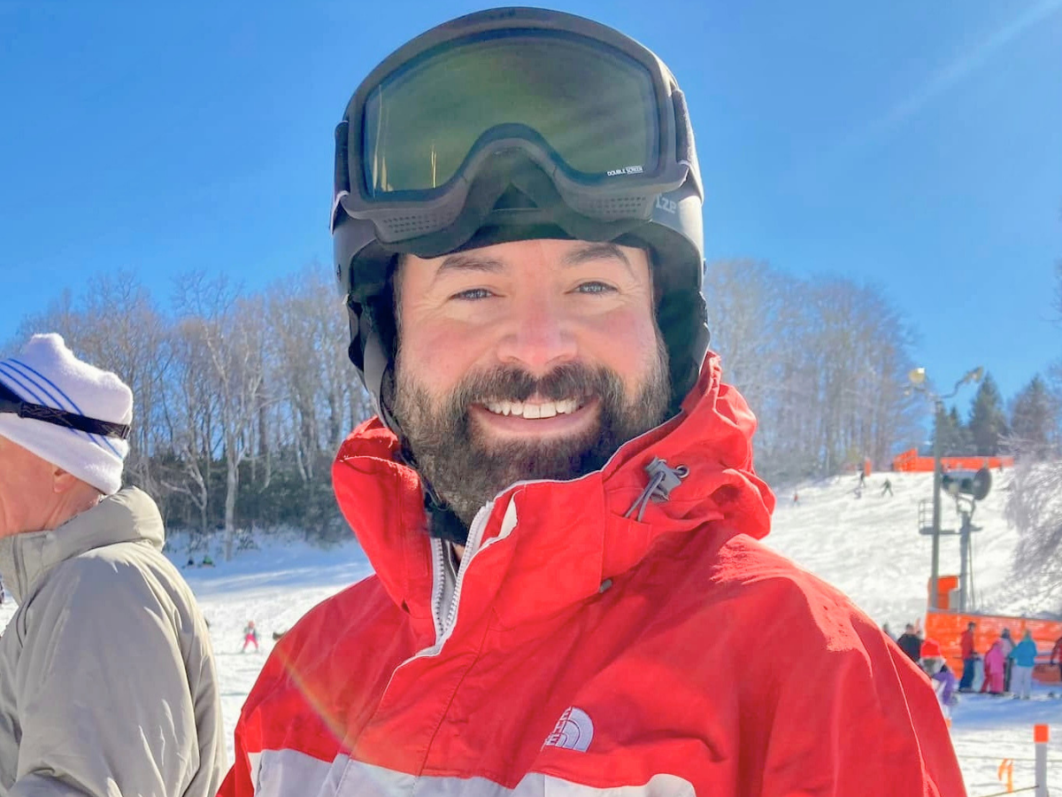 Smiling man in a red ski jacket with helmet and goggles on a sunny snowy slope.