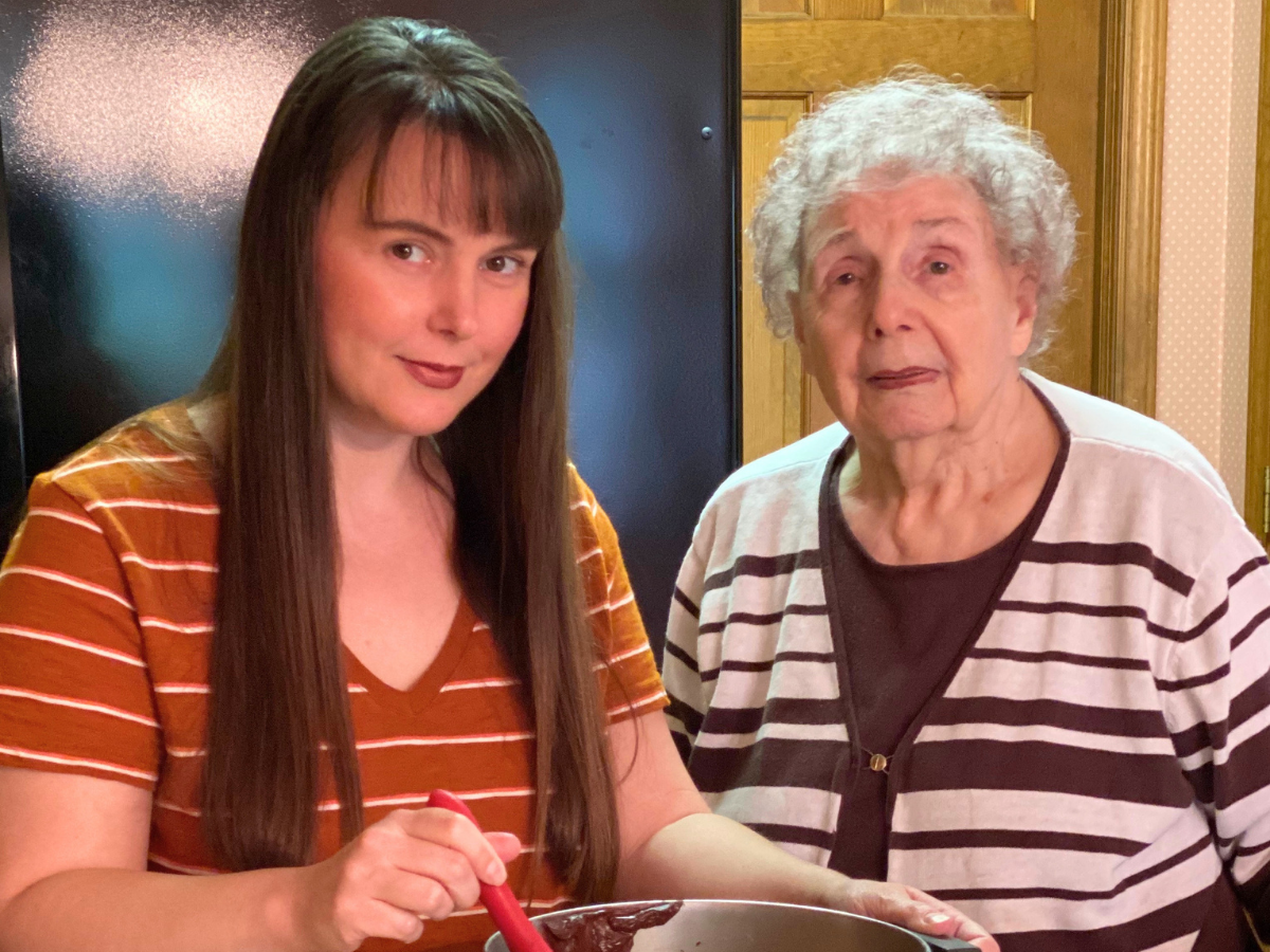 A younger woman with long brown hair in an orange striped shirt stirs batter with a red spatula beside an elderly woman in a striped sweater in a kitchen setting.