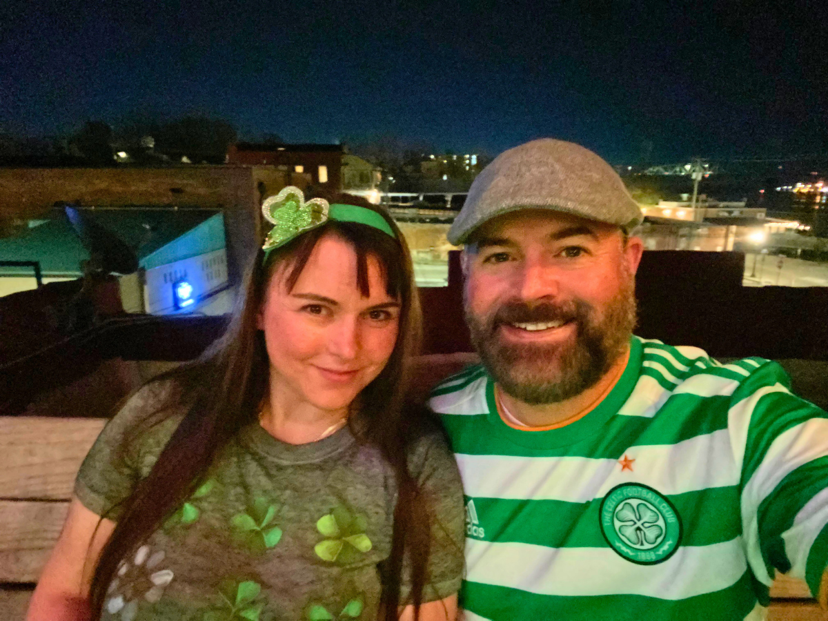 Couple taking a nighttime rooftop selfie; woman wears a shamrock headband and green shirt, man wears a green-and-white striped jersey and a flat cap, both smiling.