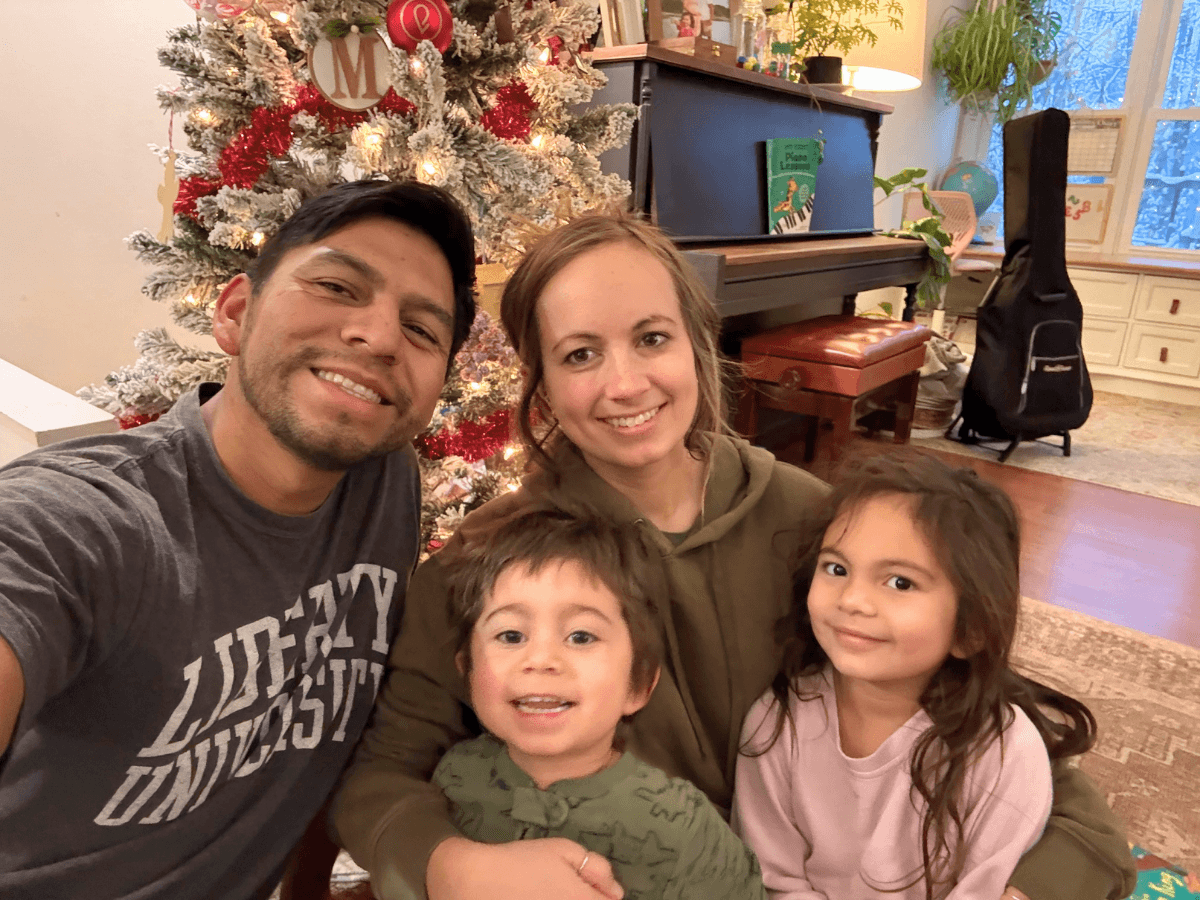 Smiling family of four takes a selfie in a living room by a lit Christmas tree, with a piano and guitar case in the background.