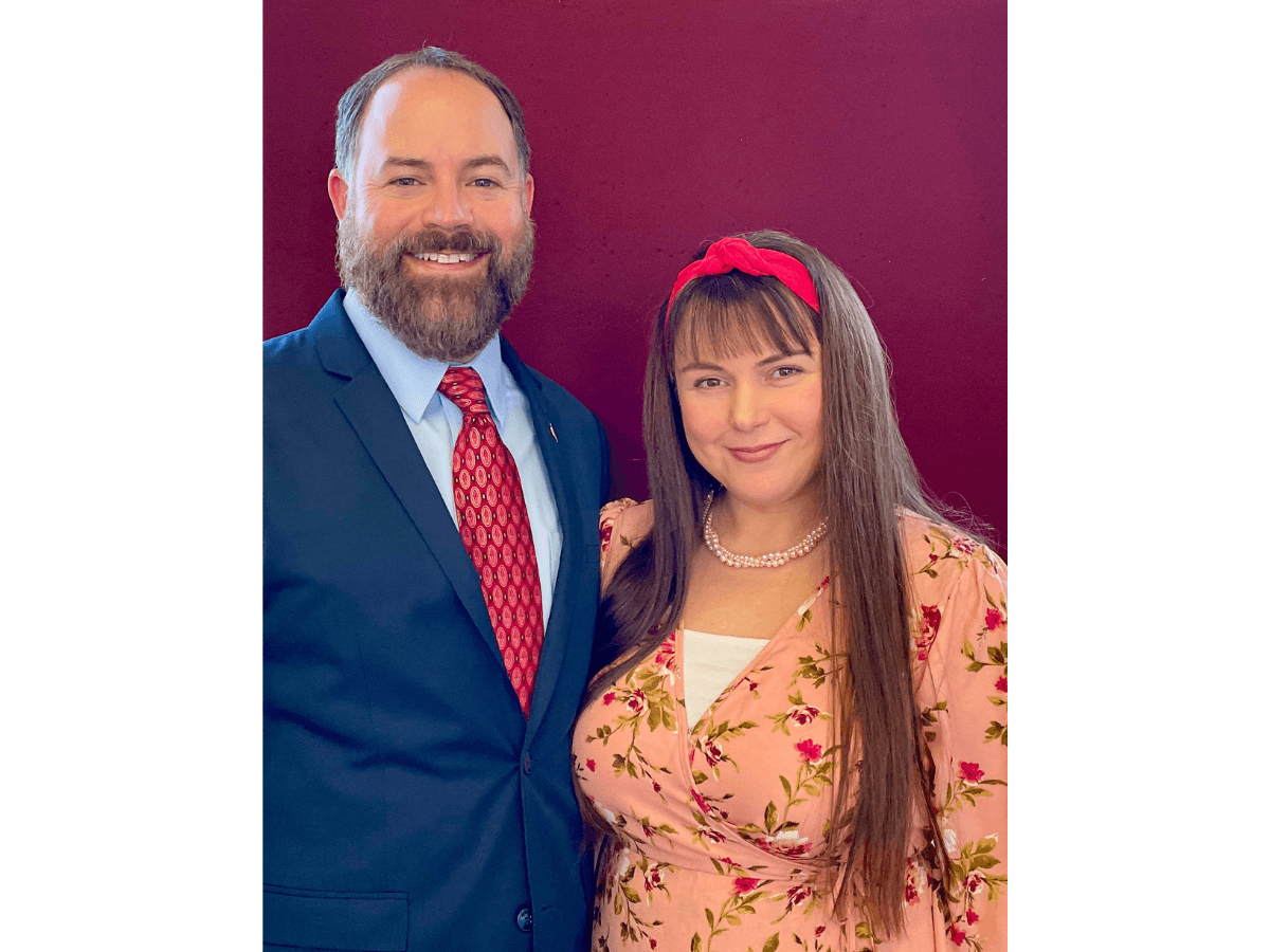 Smiling couple posing together: man in navy suit and red tie, woman in pink floral dress with a red headband and pearl necklace.