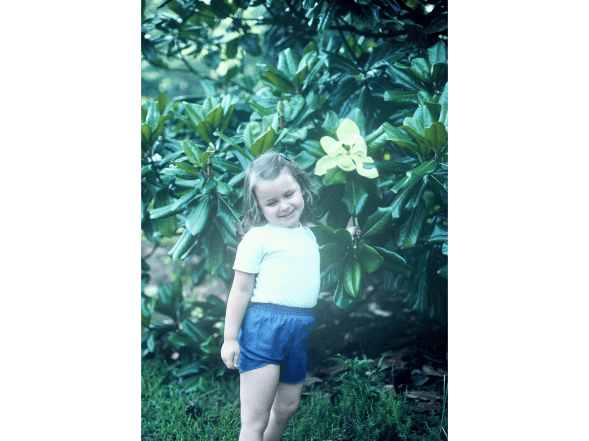 Little girl standing outdoors by green bushes with a large yellow flower, smiling, in a white shirt and blue shorts.