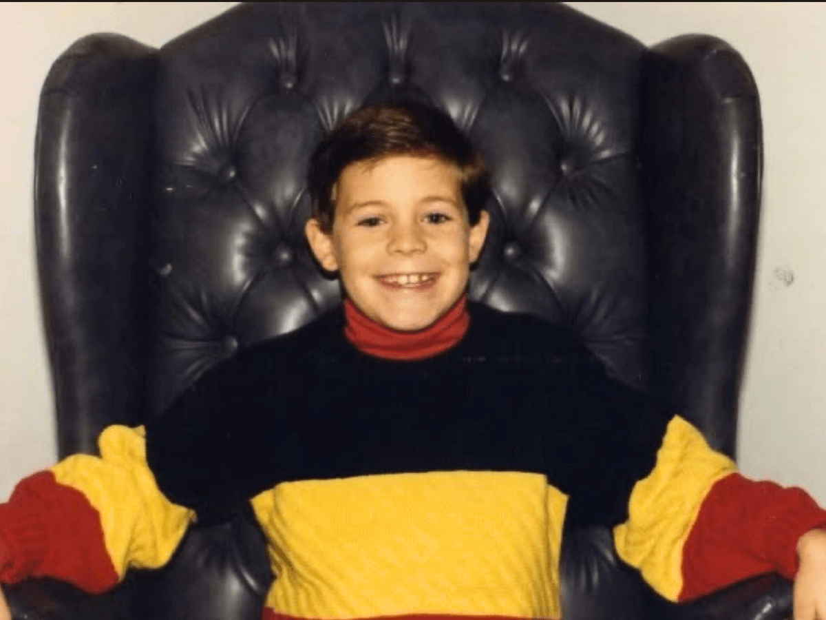 Smiling boy in a red turtle neck and oversized yellow-black outfit sits in a tall, black tufted chair.