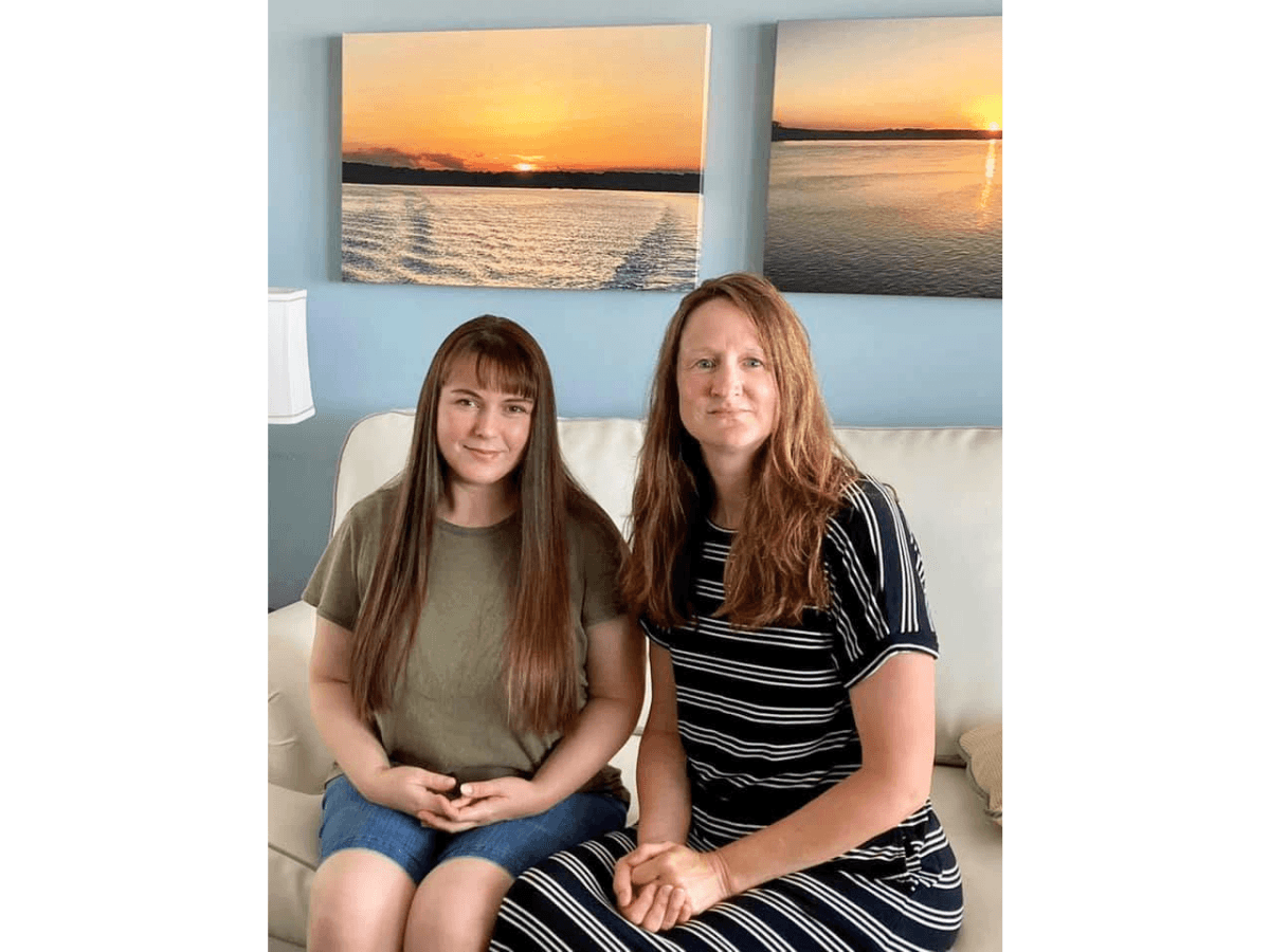 Two people sit side by side on a white sofa in a living room with blue walls and sunset wall art behind them, smiling at the camera.