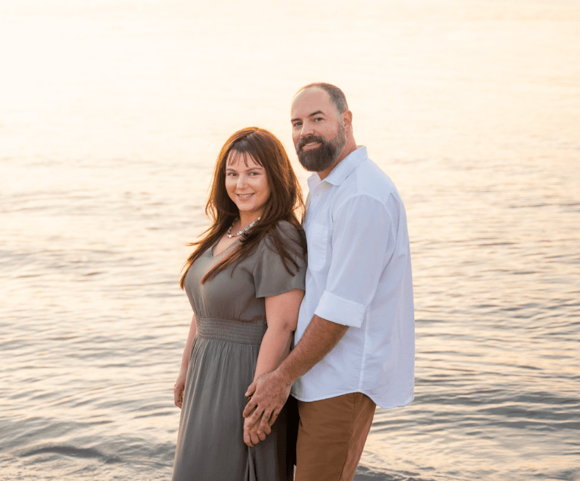 Couple standing in shallow water at sunset, holding hands and smiling by the lake.