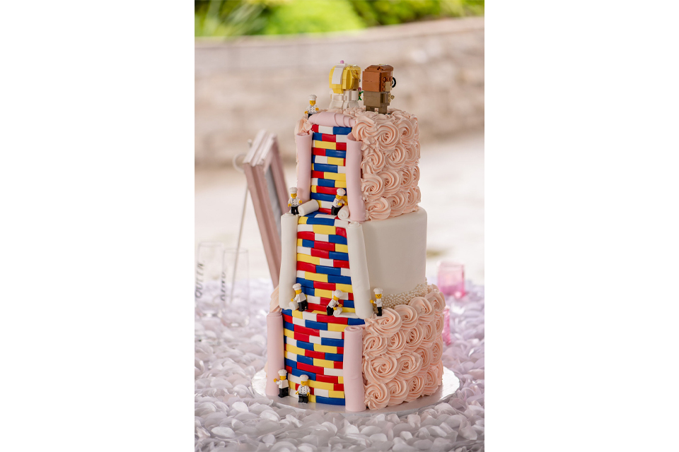 Three-tier wedding cake with pink rosette icing and colorful LEGO brick panels, topped by small LEGO figures.