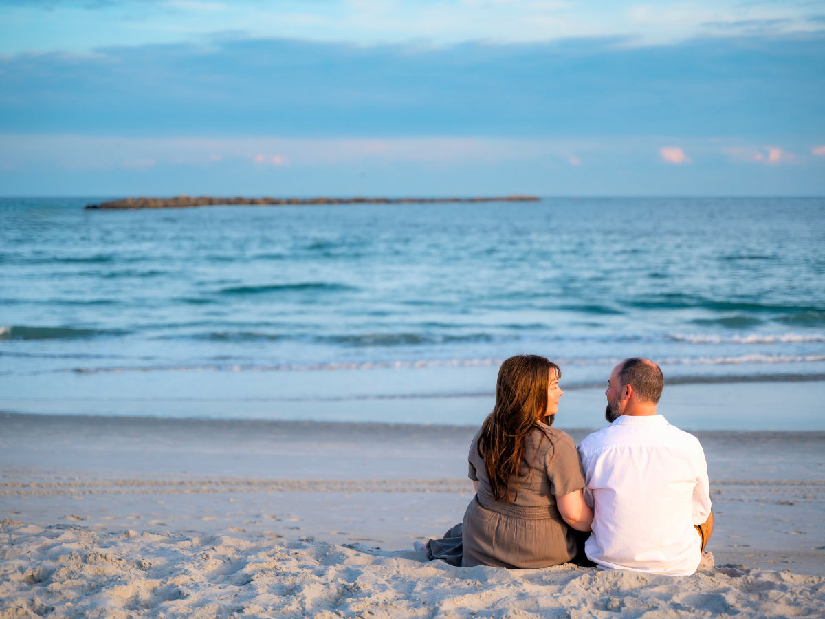Couple sitting on the sandy beach facing the calm sea, holding hands and sharing a moment as waves roll in. In the distance a rocky jetty stretches across the water.