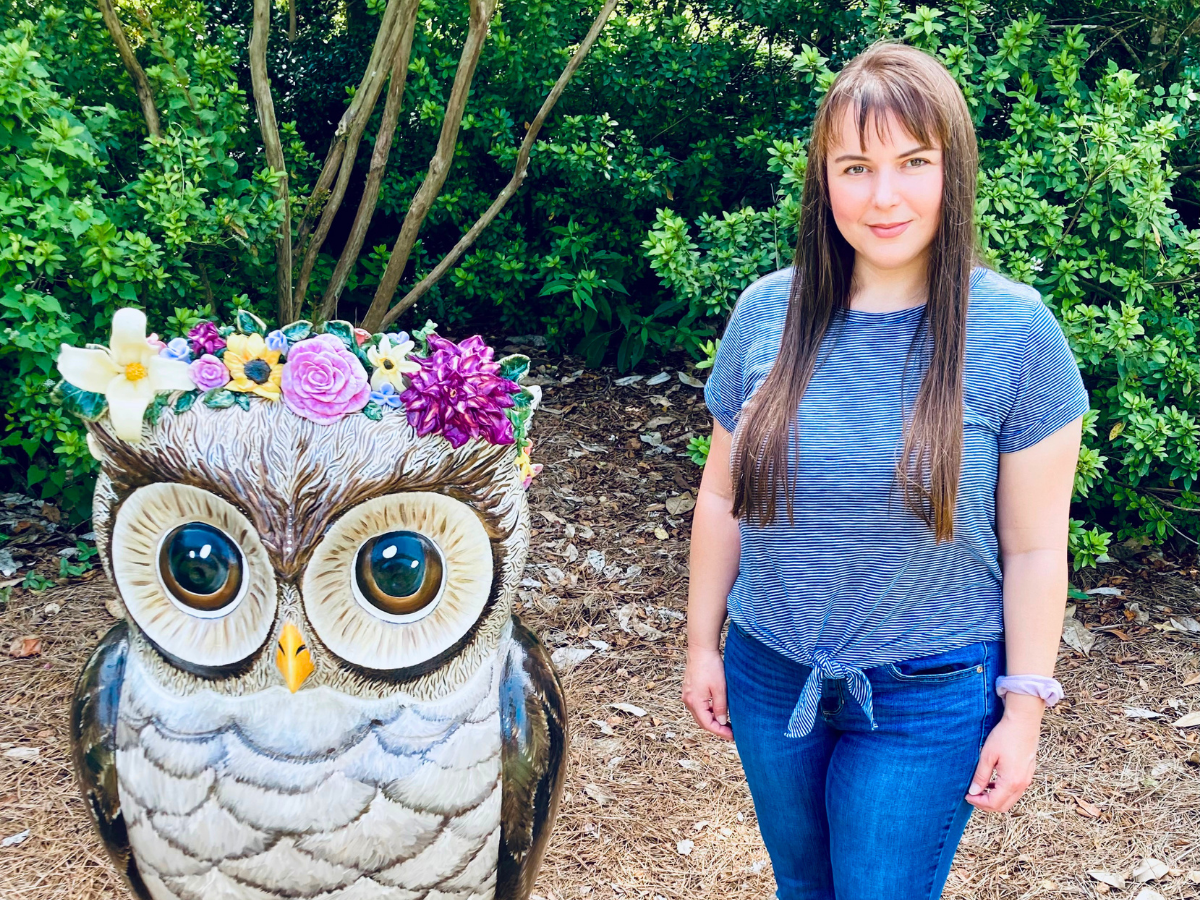 Woman in a blue striped shirt poses next to a large owl sculpture wearing a colorful flower crown in a garden setting