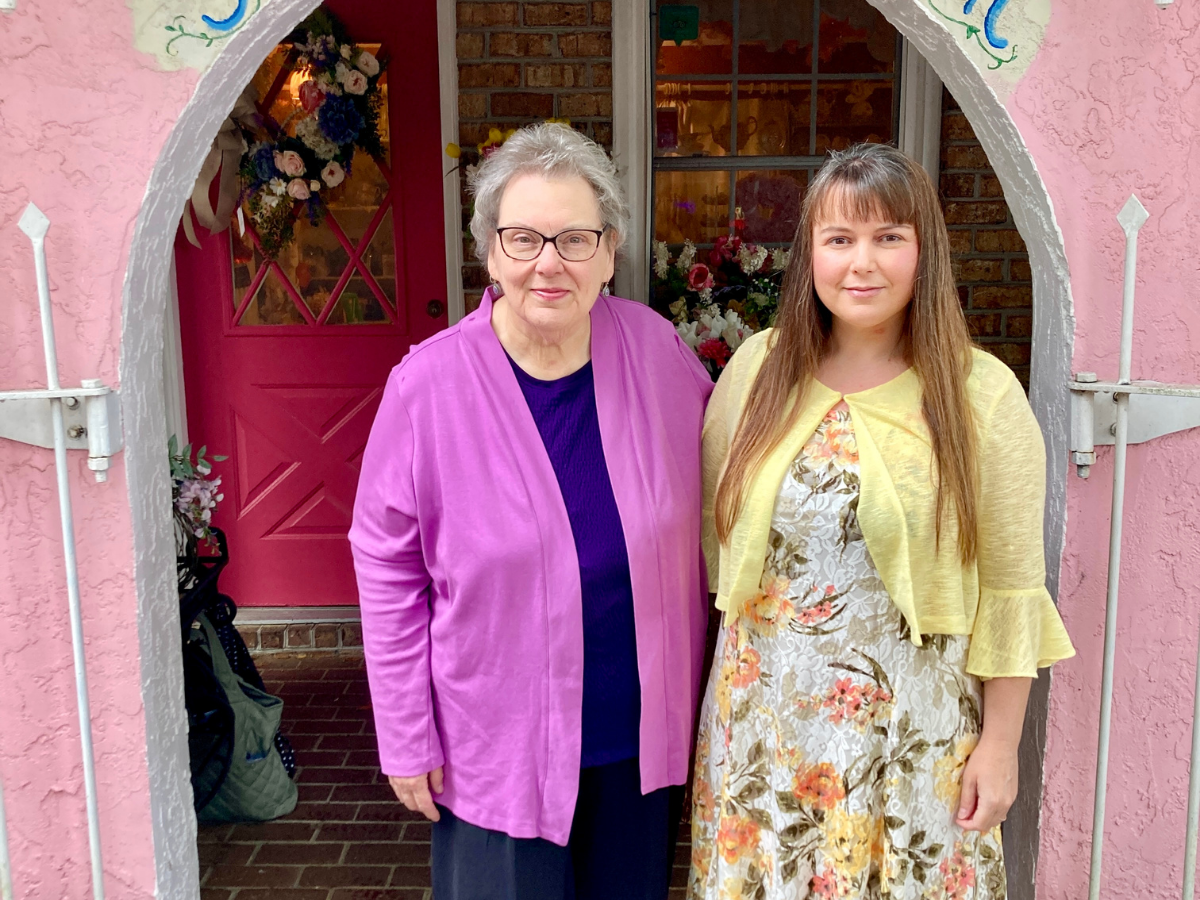 Two women pose together in a pink doorway, one older in a purple cardigan and the other in a floral dress with a yellow cardigan pale backdrop.
