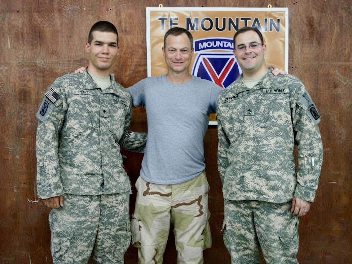 Three men pose together in front of a banner; two soldiers in camouflage uniforms flank a man in a gray t-shirt and beige camouflage shorts.