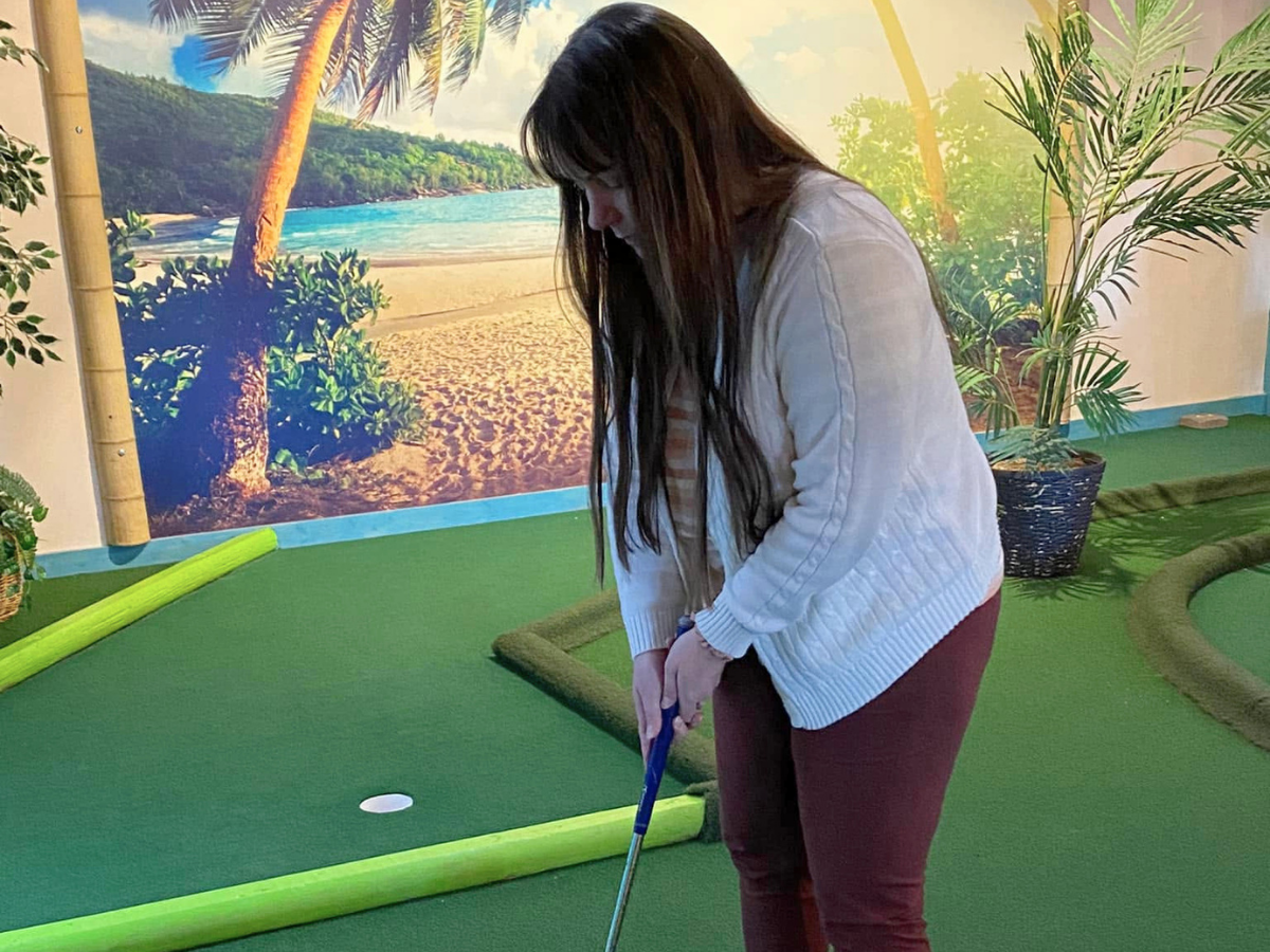 Woman in a white sweater and burgundy pants lining up a putt on an indoor mini-golf course, with a tropical mural in the background.