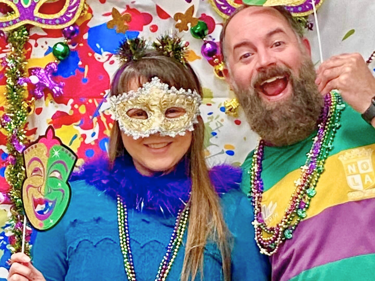 Two adults at a festive Mardi Gras scene smiling for the camera; the woman wears a gold ornamental mask and blue outfit, the man wears a green and purple shirt with many bead necklaces, colorful background with confetti and masks.