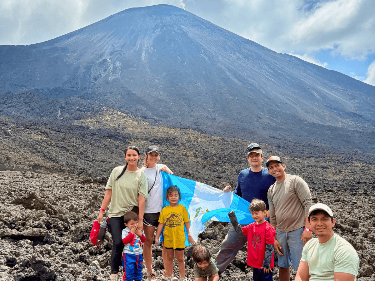 Group of adults and children posing on rough volcanic rock with a blue flag, volcano in background.