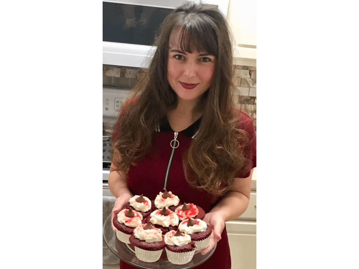 Smiling woman in a red dress holds a plate of cupcakes with white frosting and red toppings on top.