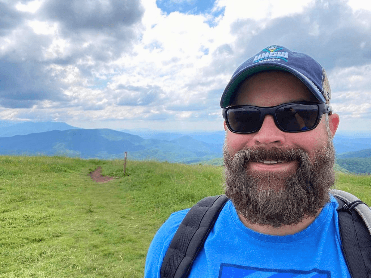 Man with beard wearing sunglasses and a blue shirt, hiking on a grassy hill with distant blue mountains and a cloudy sky in the background, backpack visible.