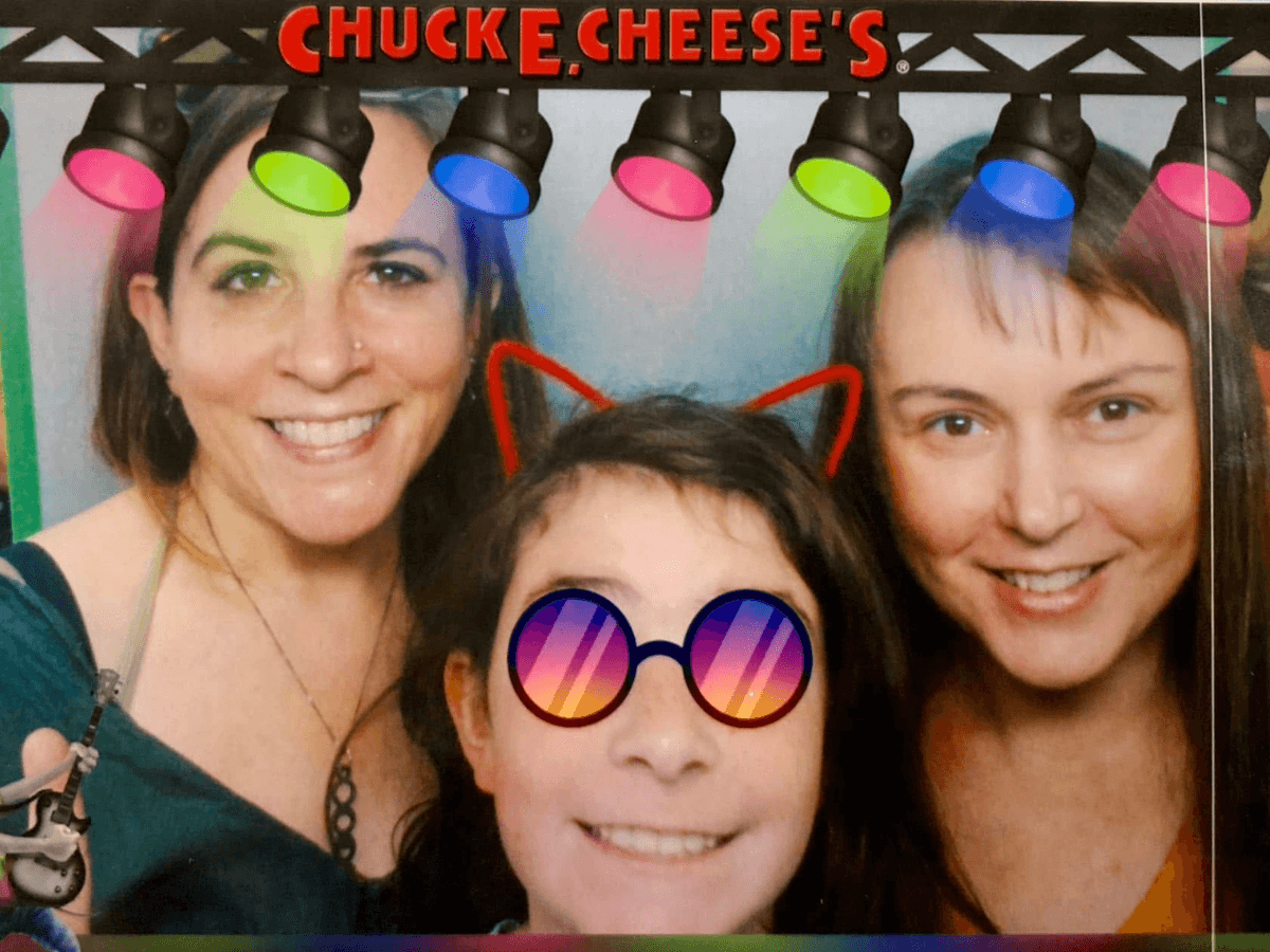 Three smiling people pose in a Chuck E. Cheese's photo booth; center child wears rainbow sunglasses and a red cat-ear headband.