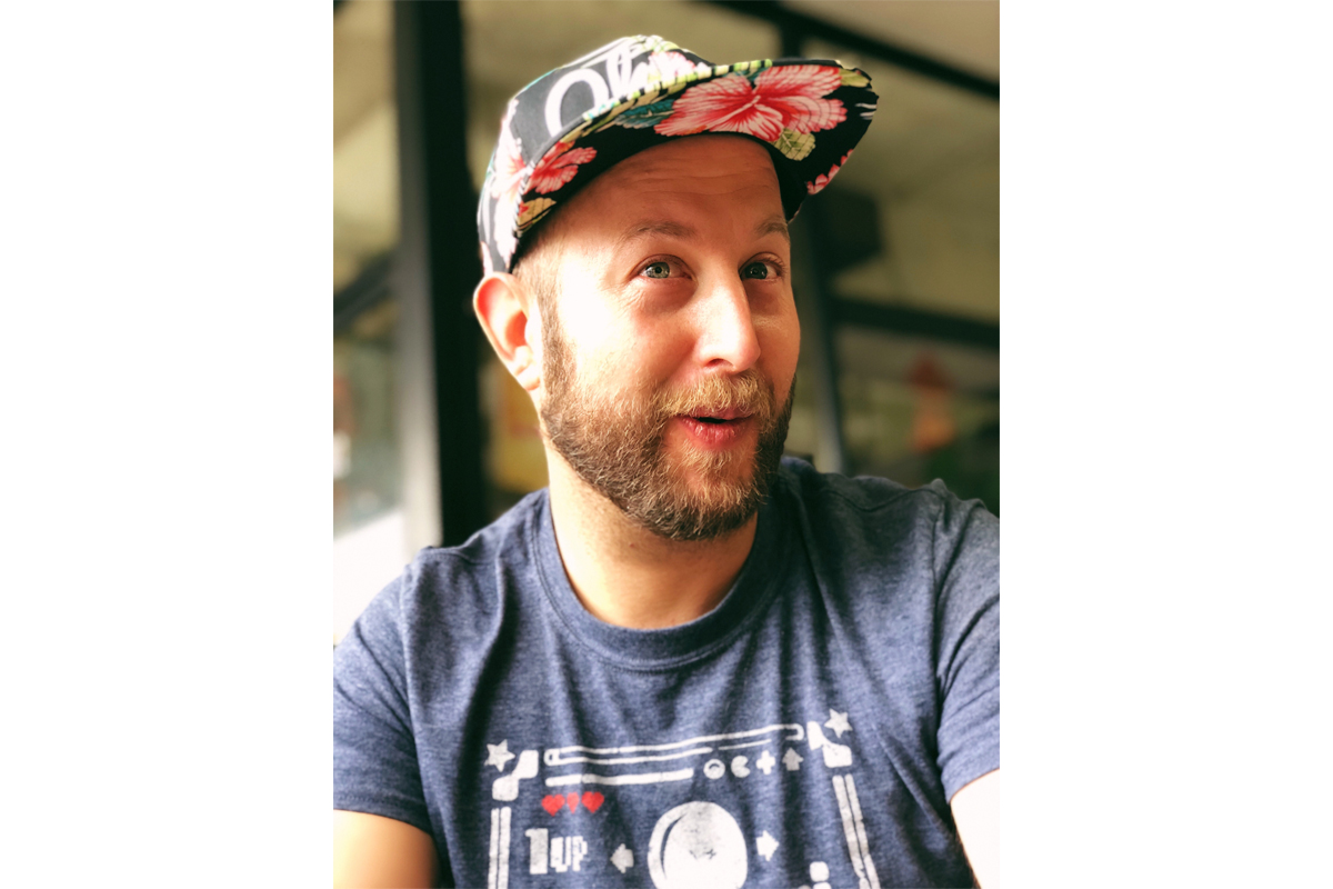 Man with a beard wearing a tropical floral cap and blue T-shirt, looking off to the side, in a casual outdoor setting