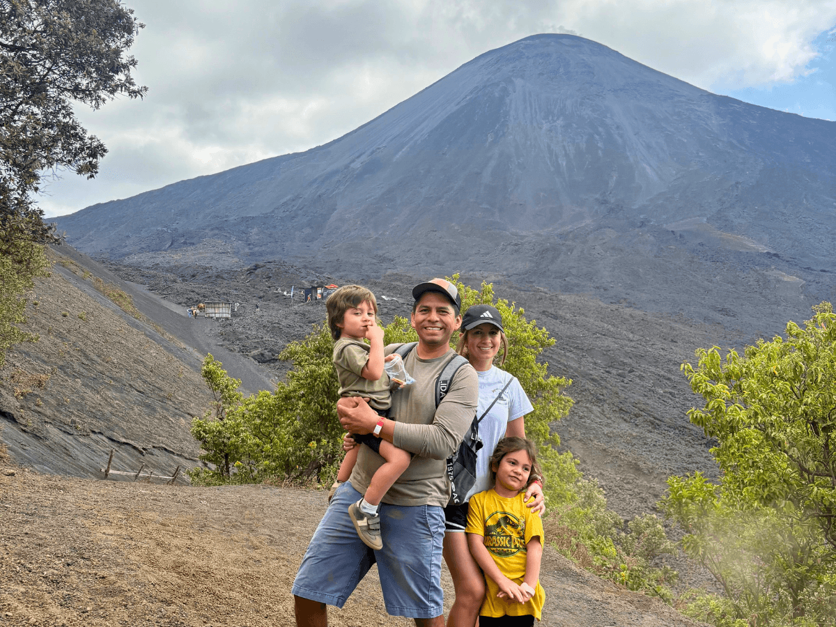 Family of four posing in front of a large, dark volcanic cone and lava field behind them to the right side of the frame.