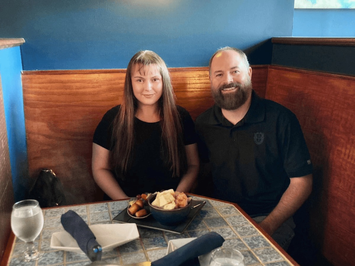 A couple sitting in a restaurant booth with a bowl of chips and dip on the table, smiling at the camera.