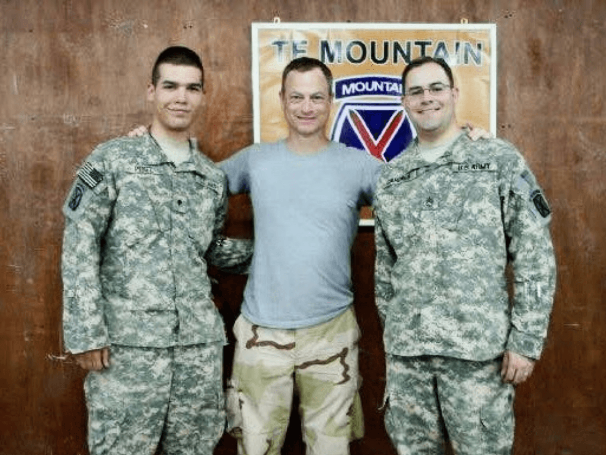 Three men pose with two soldiers in camouflage uniforms flanking a civilian in a gray t‑shirt in front of a 'The Mountain' sign.