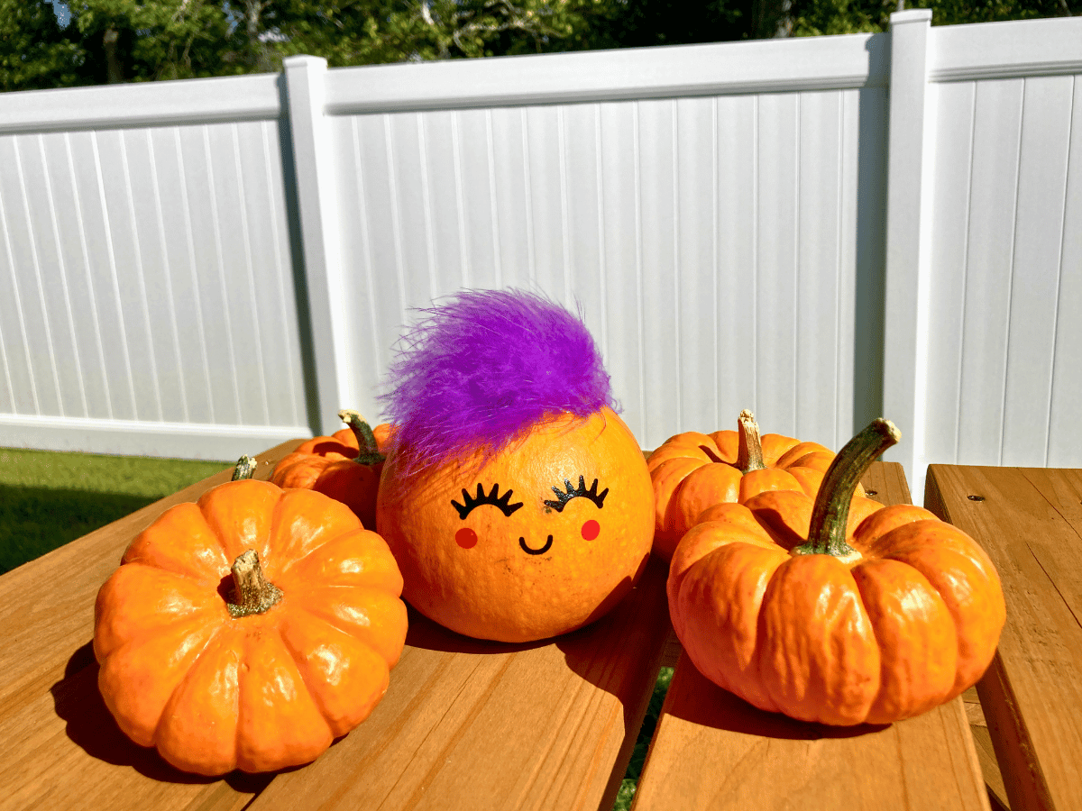 Three pumpkins on a wooden outdoor table; the middle pumpkin has a purple feather and a smiling face drawn on it.