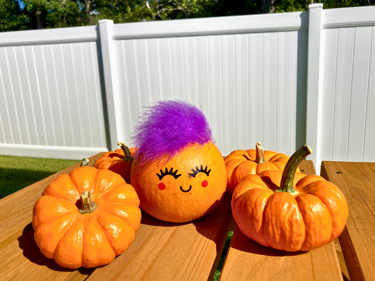 Three orange pumpkins on a wooden table outdoors, with the center pumpkin sporting purple fur and a cute drawn face.