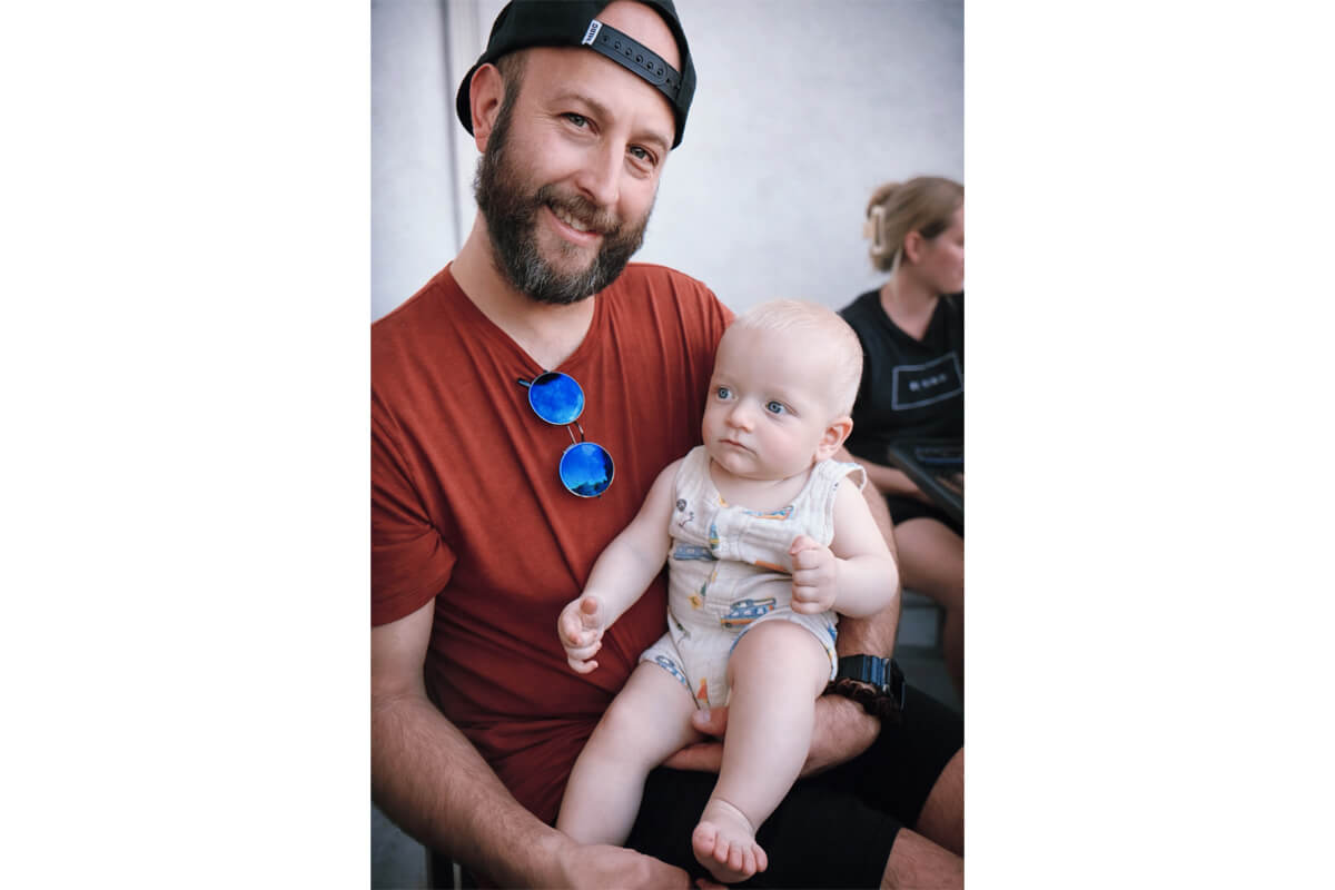Father with a beard and backward cap holds a baby on his lap; blue sunglasses hang from his shirt.