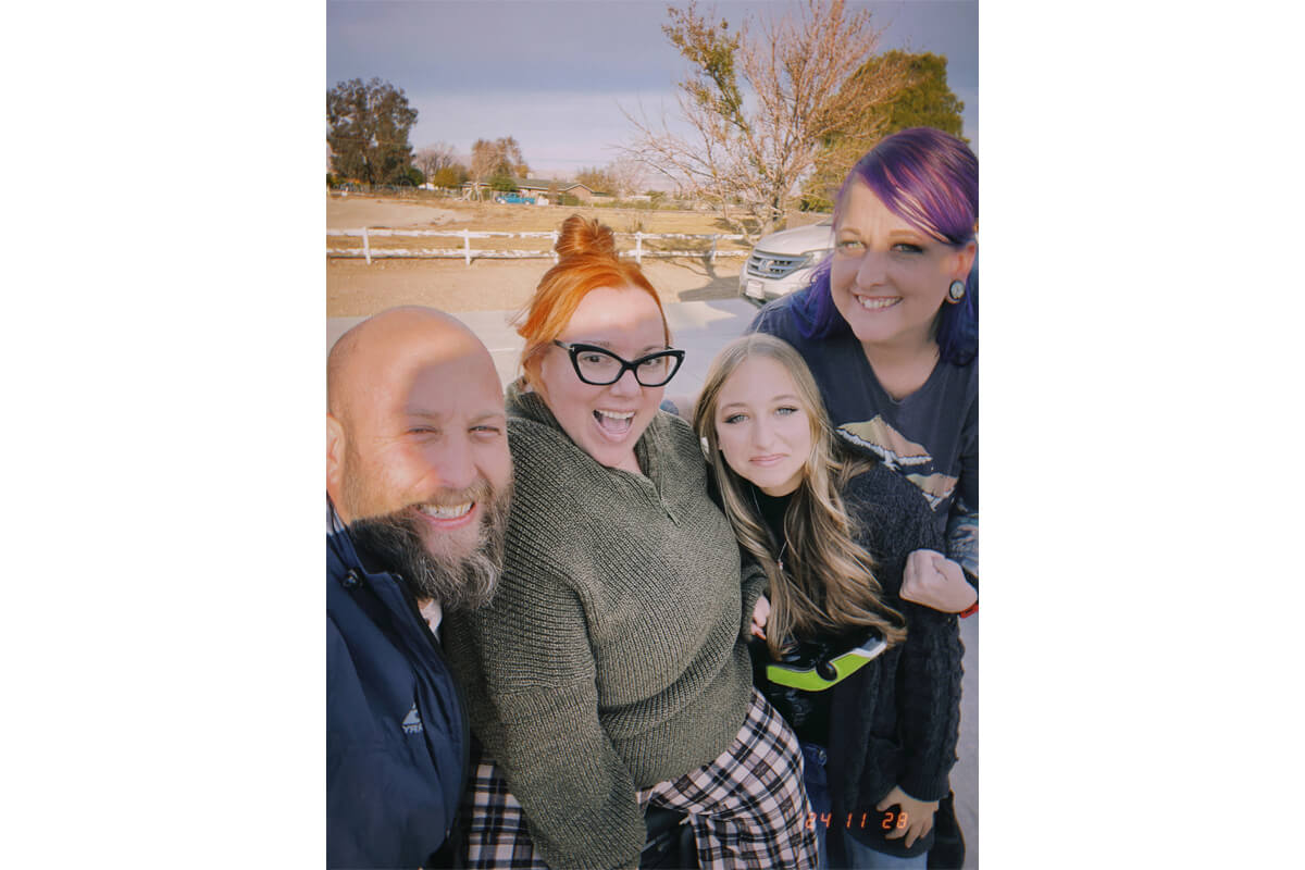 Four people smile for a selfie outdoors, standing close together with a white fence and bare trees in the background. (Group of four posing outside on a sunny day.)