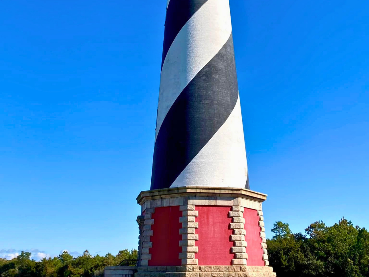 Lighthouse with black-and-white diagonal stripes on a red brick base, set against a clear blue sky and green trees.