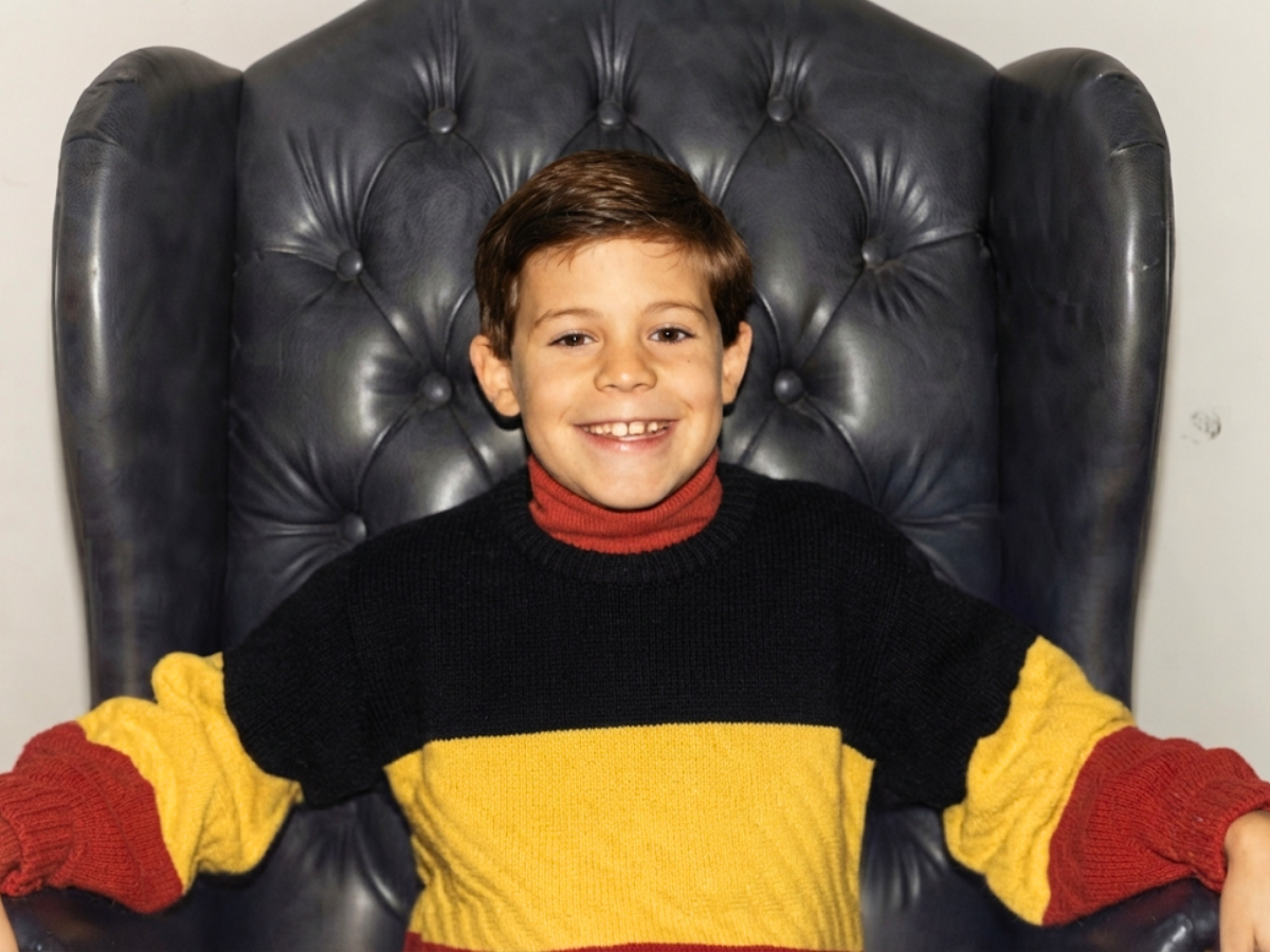 Smiling boy sits in a tall dark leather chair, wearing a red turtleneck and a yellow-black-red color-block sweater.