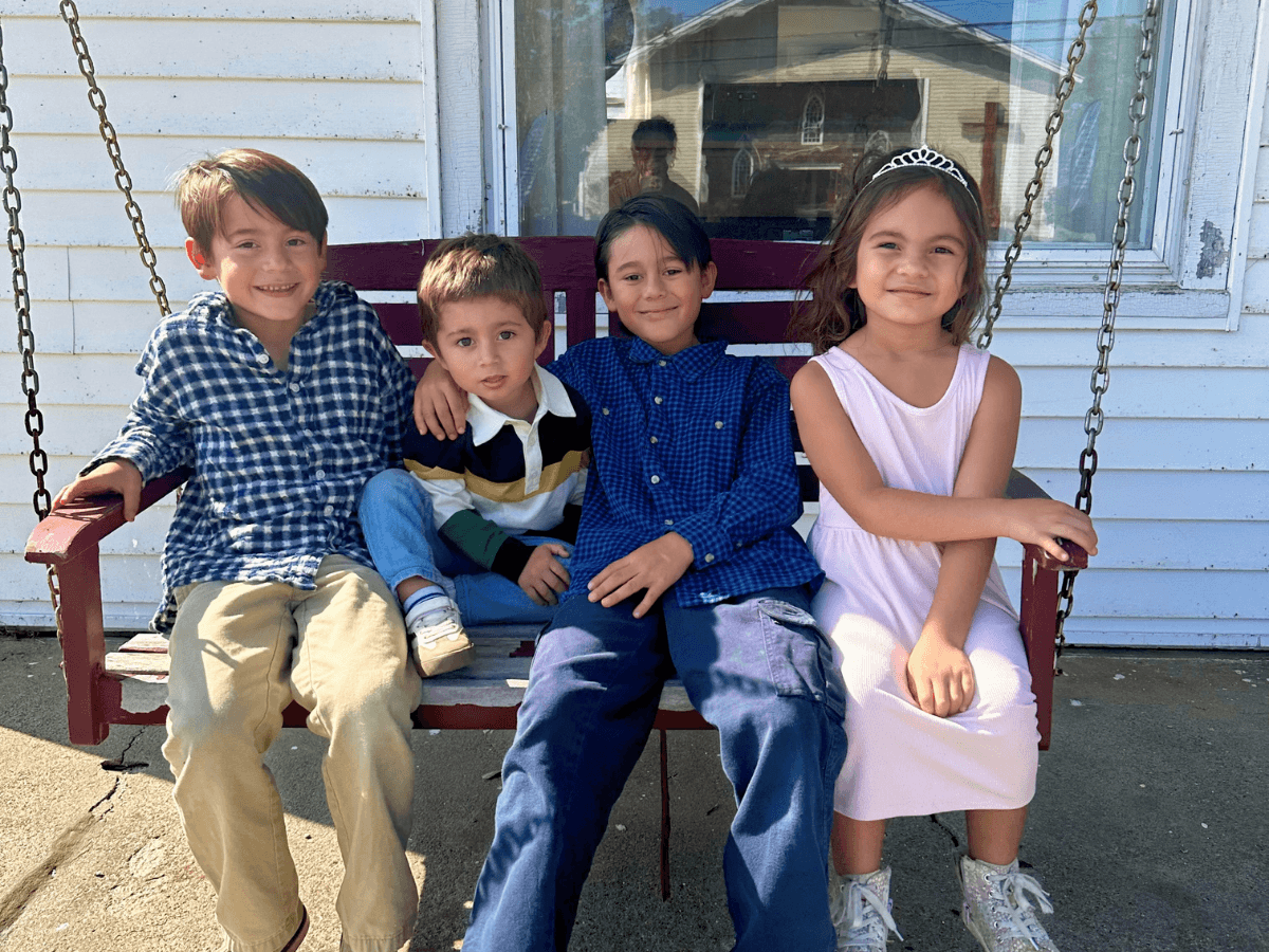 Four smiling children sit on a wooden swing outside a white house; two boys on the left, a girl in a pink dress with a tiara on the right.